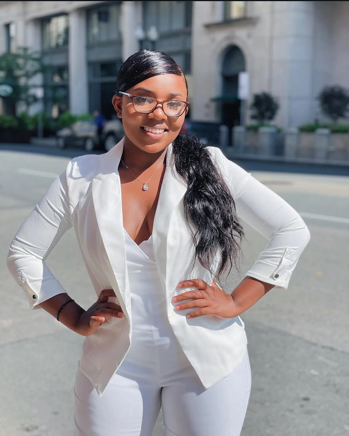 Woman with glasses and long black hair standing outdoors on a city street, wearing a white blazer and white pants, smiling with hands on hips.