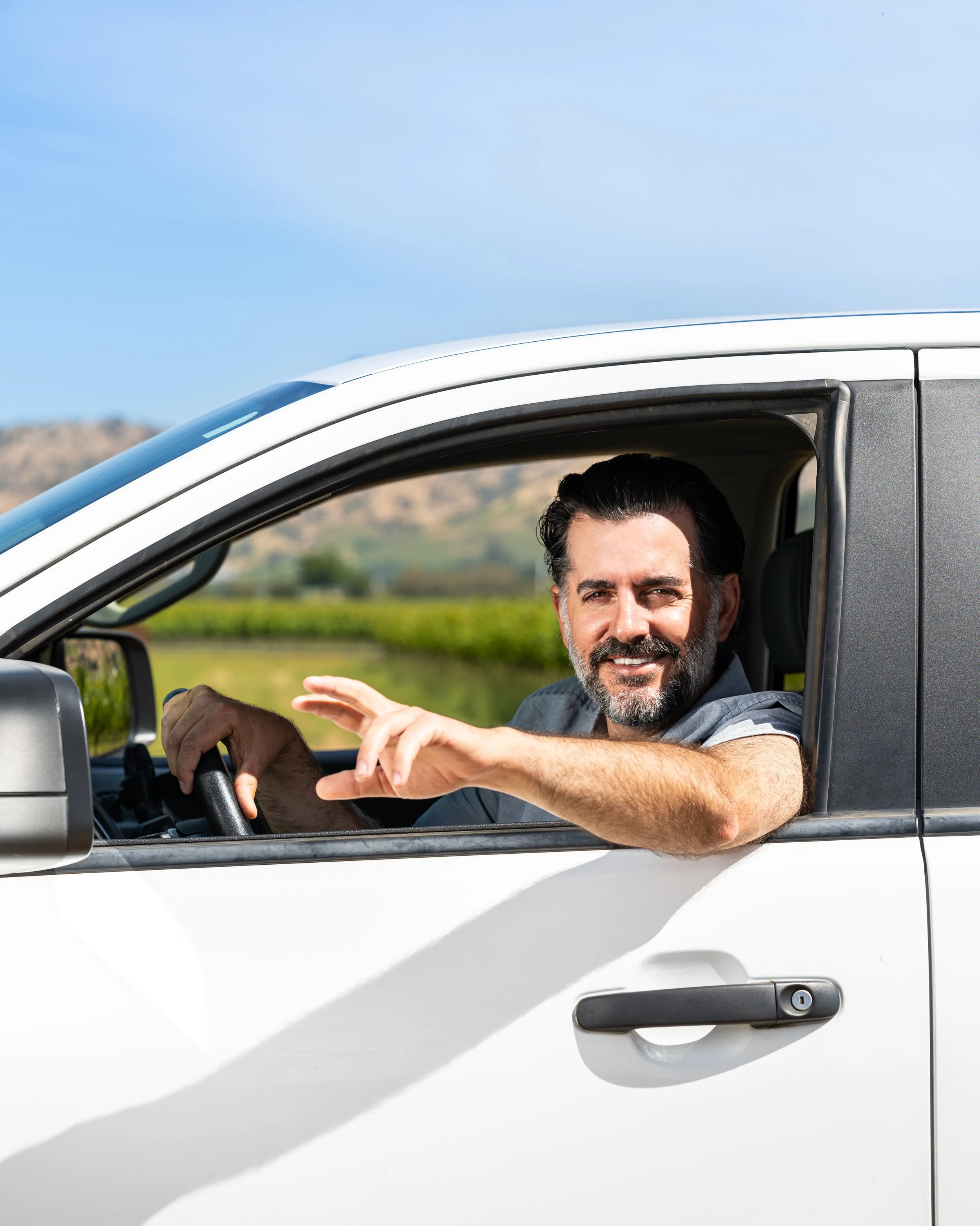 Smiling man with dark hair and beard driving a white vehicle in a rural area under a blue sky.
