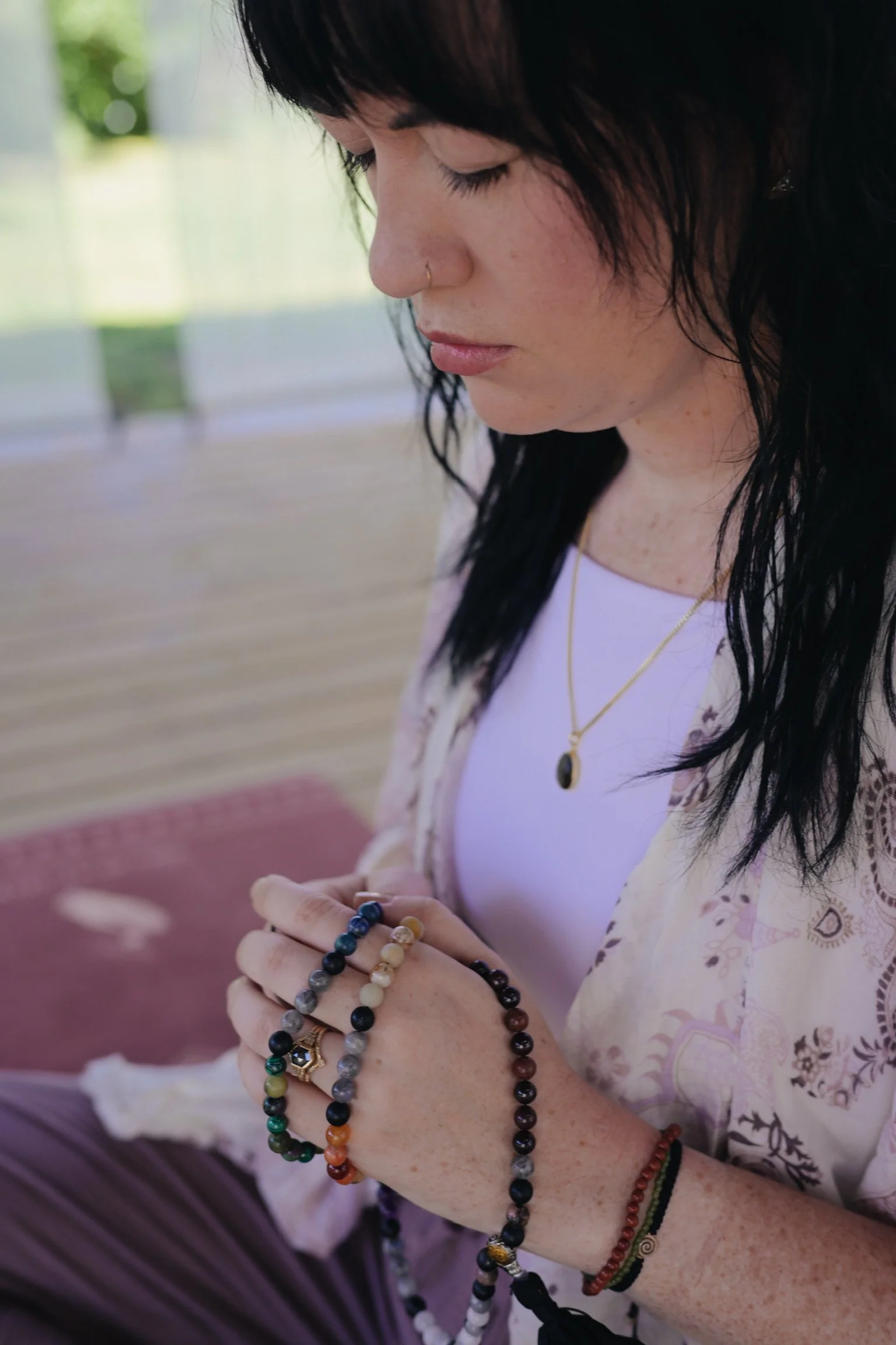 A woman with dark hair and freckles is sitting outdoors with a blurred green background, wearing multiple bracelets on her wrists and holding her hands together.