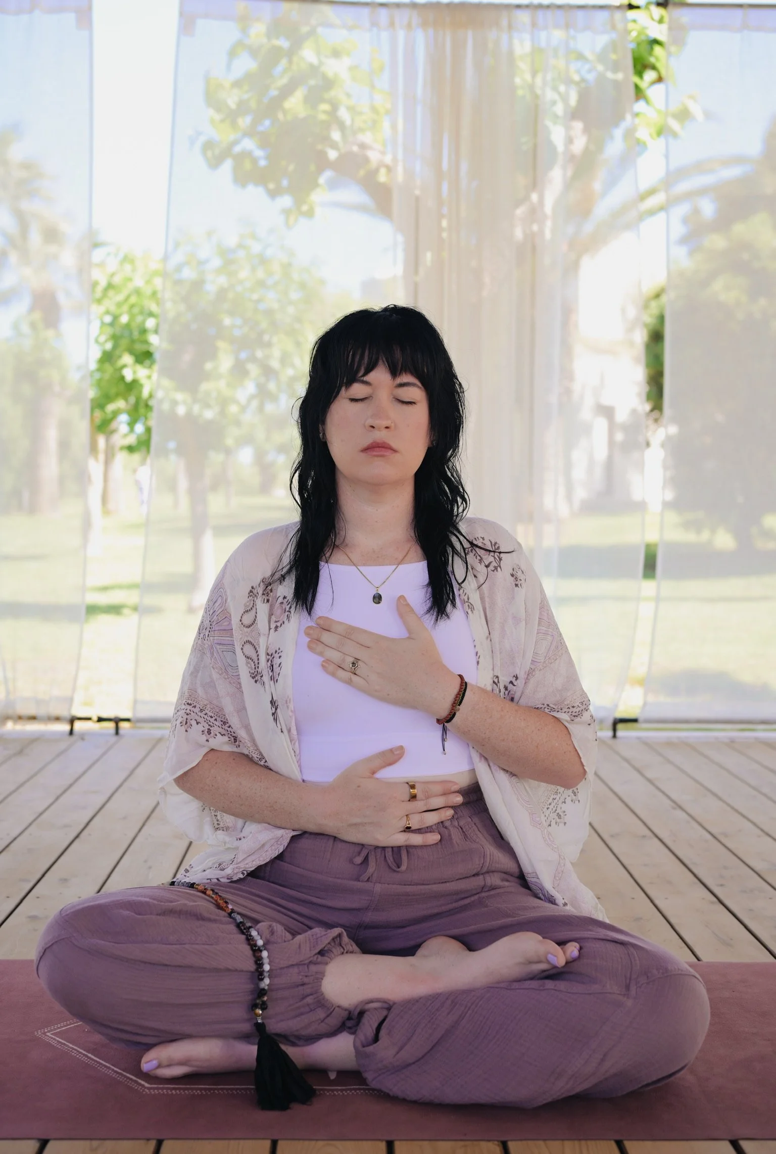 A woman practicing yoga outdoors on a pink yoga mat, sitting cross-legged with eyes closed, hands on her chest, in front of large glass windows with trees visible outside.