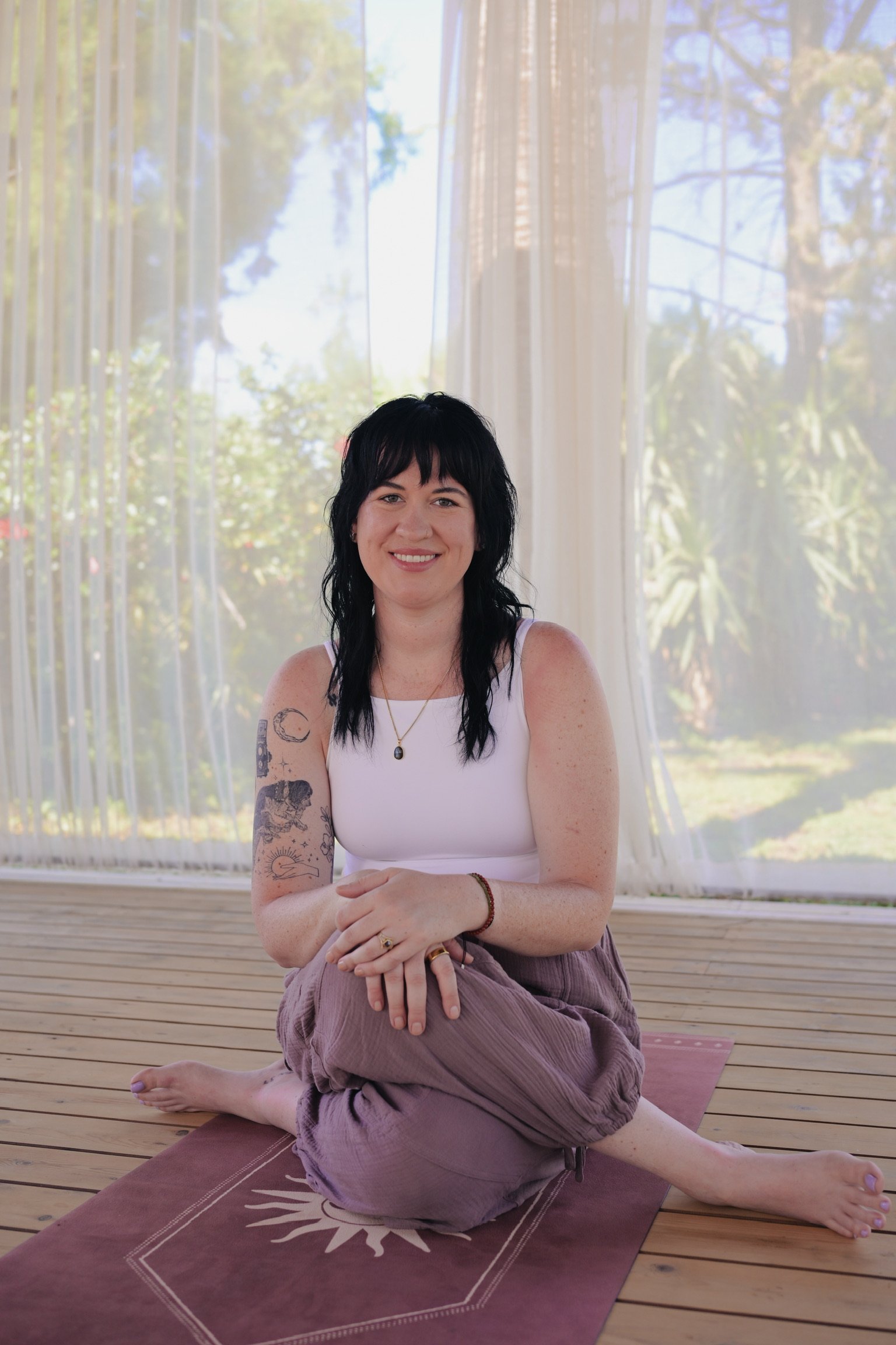 A woman with black hair and tattoos on her arms sitting on a yoga mat indoors, practicing yoga with a background of large windows showing trees and sky.