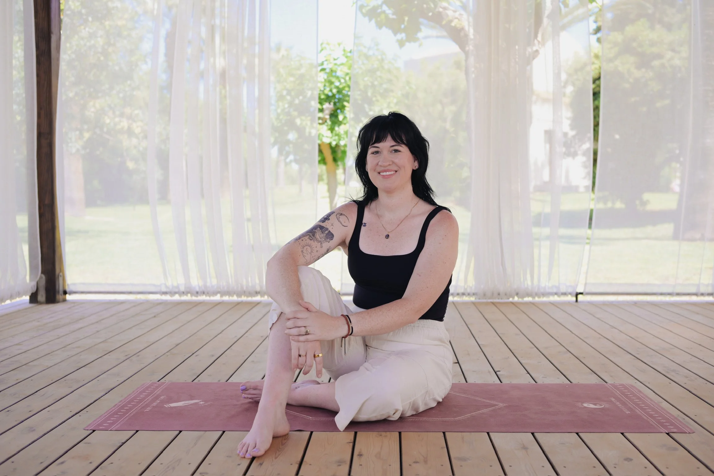 A woman with black hair, tattoos on her left arm, sitting on a yoga mat in a sunny outdoor setting with trees and white curtains in the background, smiling at the camera.