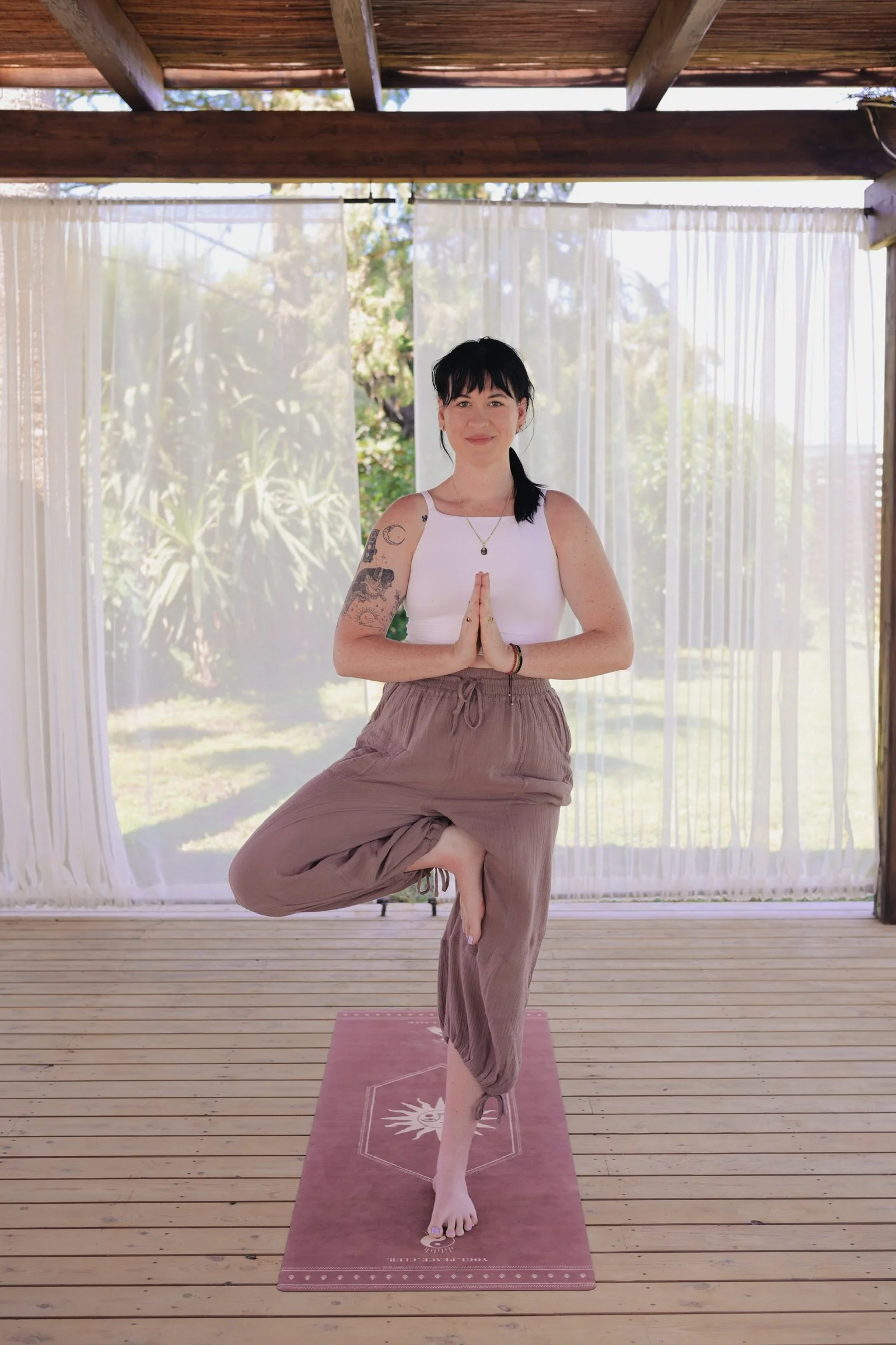 A woman practicing yoga outdoors on a wooden deck, standing on a pink yoga mat, in a tree pose with hands in prayer position, balancing on one leg, with a background of trees and a sheer curtain.