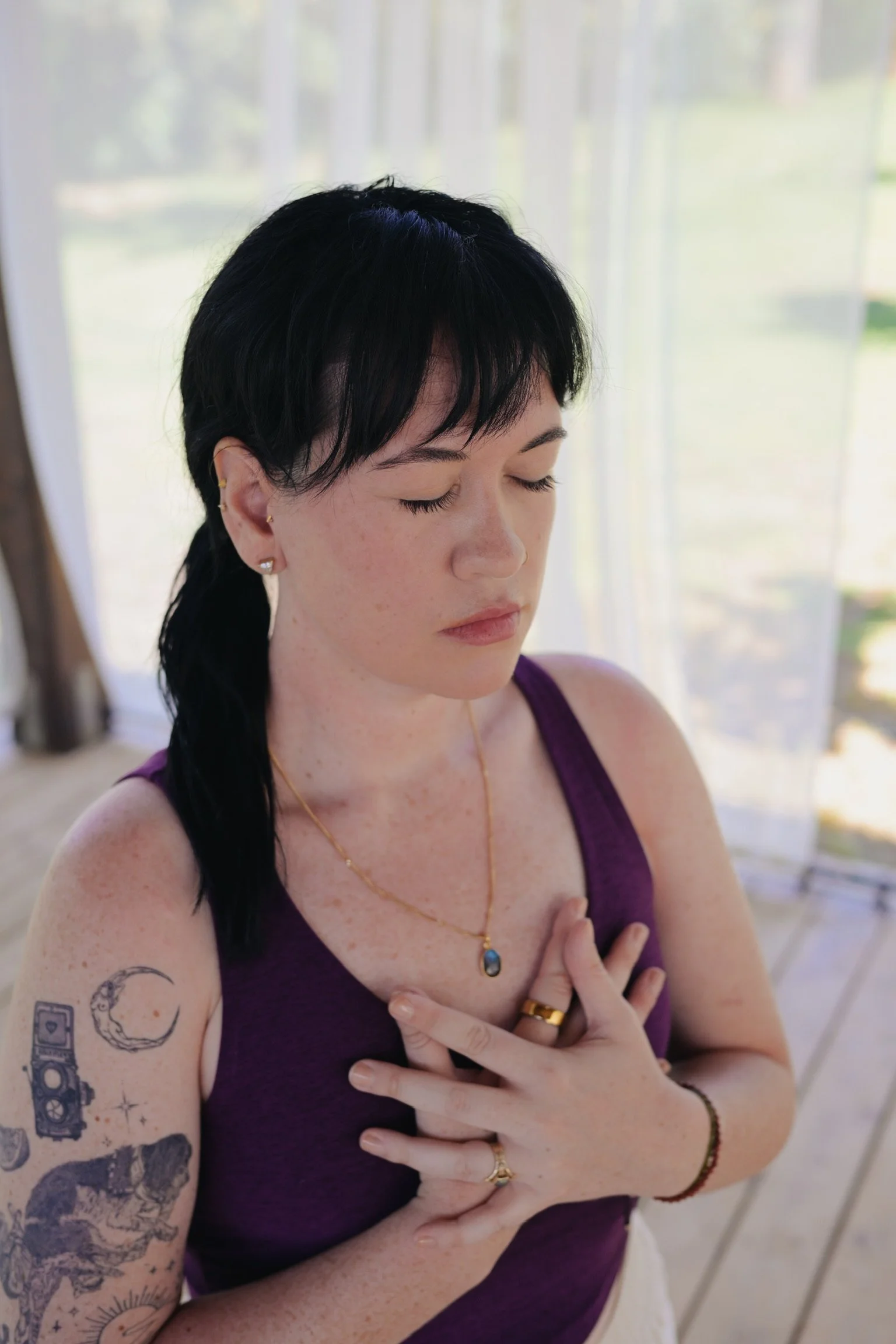 A woman with short black hair and tattoos on her left arm, wearing a purple tank top, gold jewelry, and a ring, appears to be meditating with her eyes closed, holding her hand over her chest, in front of a window with sheer curtains.