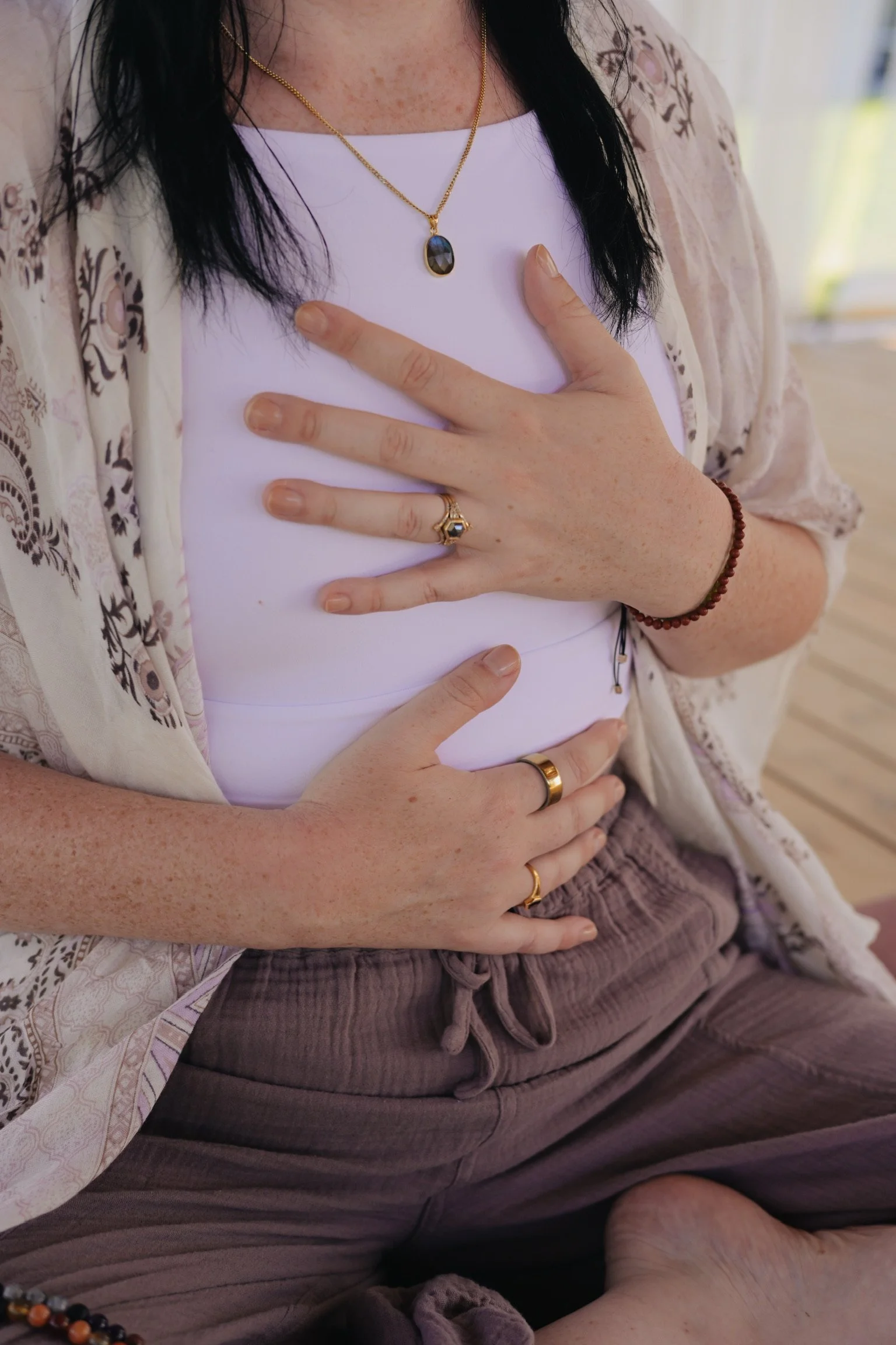 A person wearing a white shirt with a patterned beige and brown cardigan, gold necklace with a dark oval pendant, gold rings, a beaded bracelet, and sitting in a relaxed position indoors.