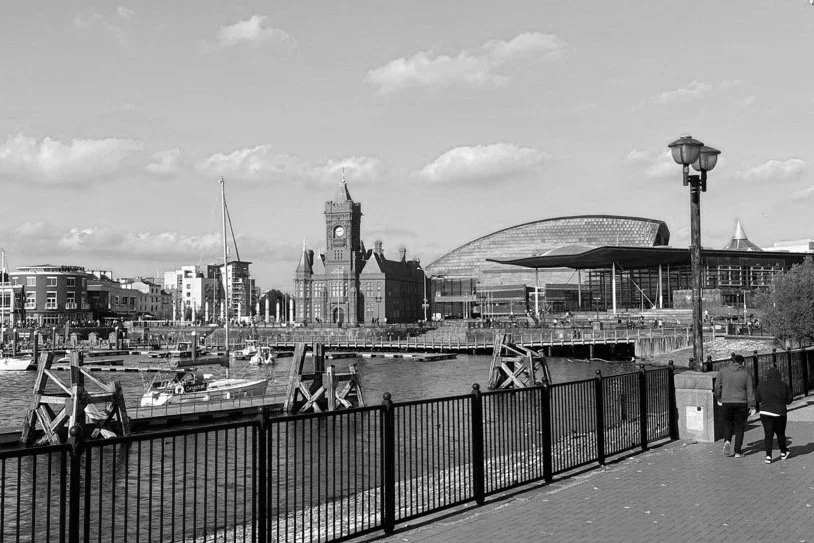 View of a waterfront with sailboats, a historic clock tower, modern buildings, a modern curved glass structure, and two people walking along a walkway with a fence and lamp post.