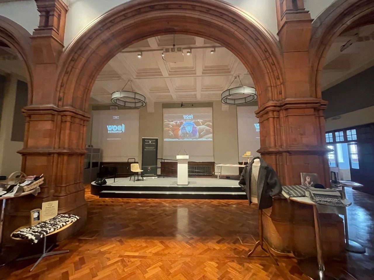 An empty stage with a white podium in the center, flanked by a chair and a laptop on a small table on the right side. A large wooden arch frames the stage, and screens behind display the word 'wool' and images of colorful yarn. The room has a wooden floor and decorative ceiling lights, with various objects and furniture scattered around.