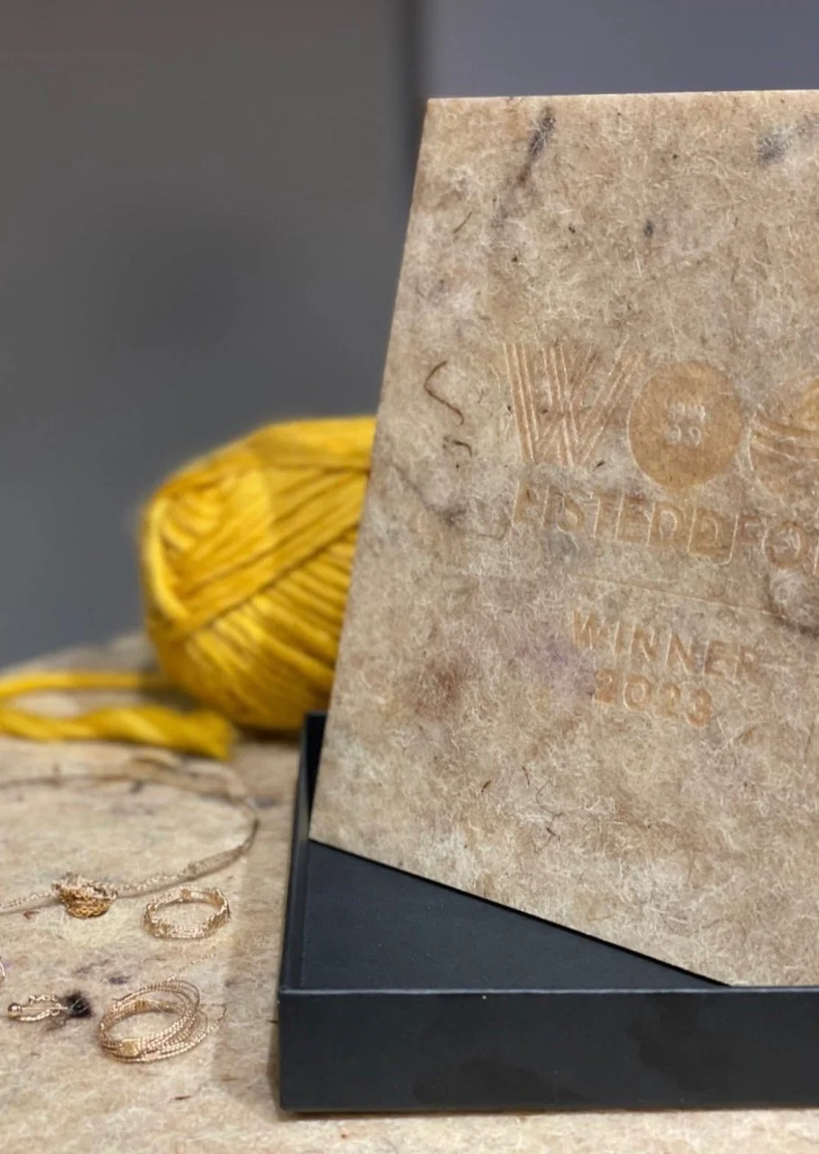 A textured beige card with faint engraved text, partially visible, leaning on a black box. Behind the card, a yellow ball of yarn and several jewelry pieces, including rings and chains, are on a surface.
