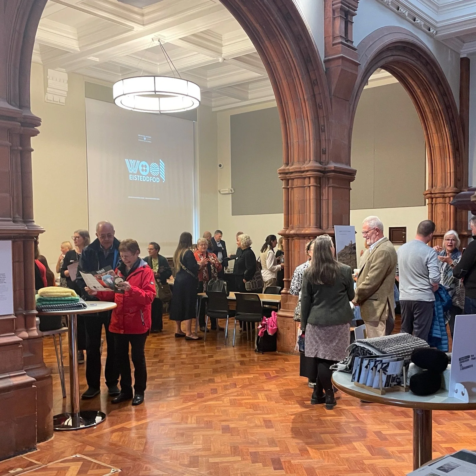 People gathered in a large, elegant room with wooden architectural features, engaging in conversations and browsing tables with various items, possibly at a conference or exhibition.