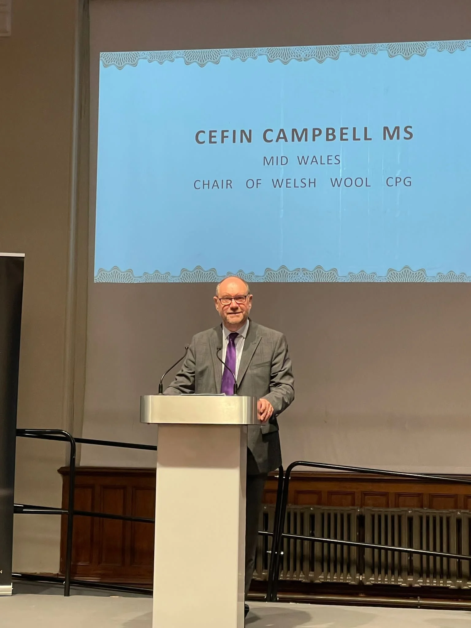 A man standing at a podium giving a presentation, with a large screen behind him displaying a slide titled 'Cefin Campbell MS’ and additional information about him.
