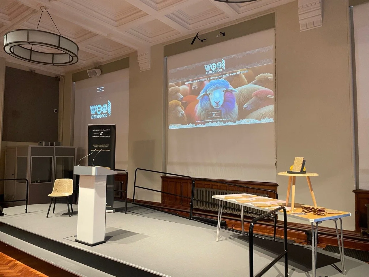 An empty conference room with a stage, a podium with microphones, a chair, a table with chairs, and a large projection screen displaying the logo "Wool Eisteddfod" with sheep images and is set up for a presentation or event.