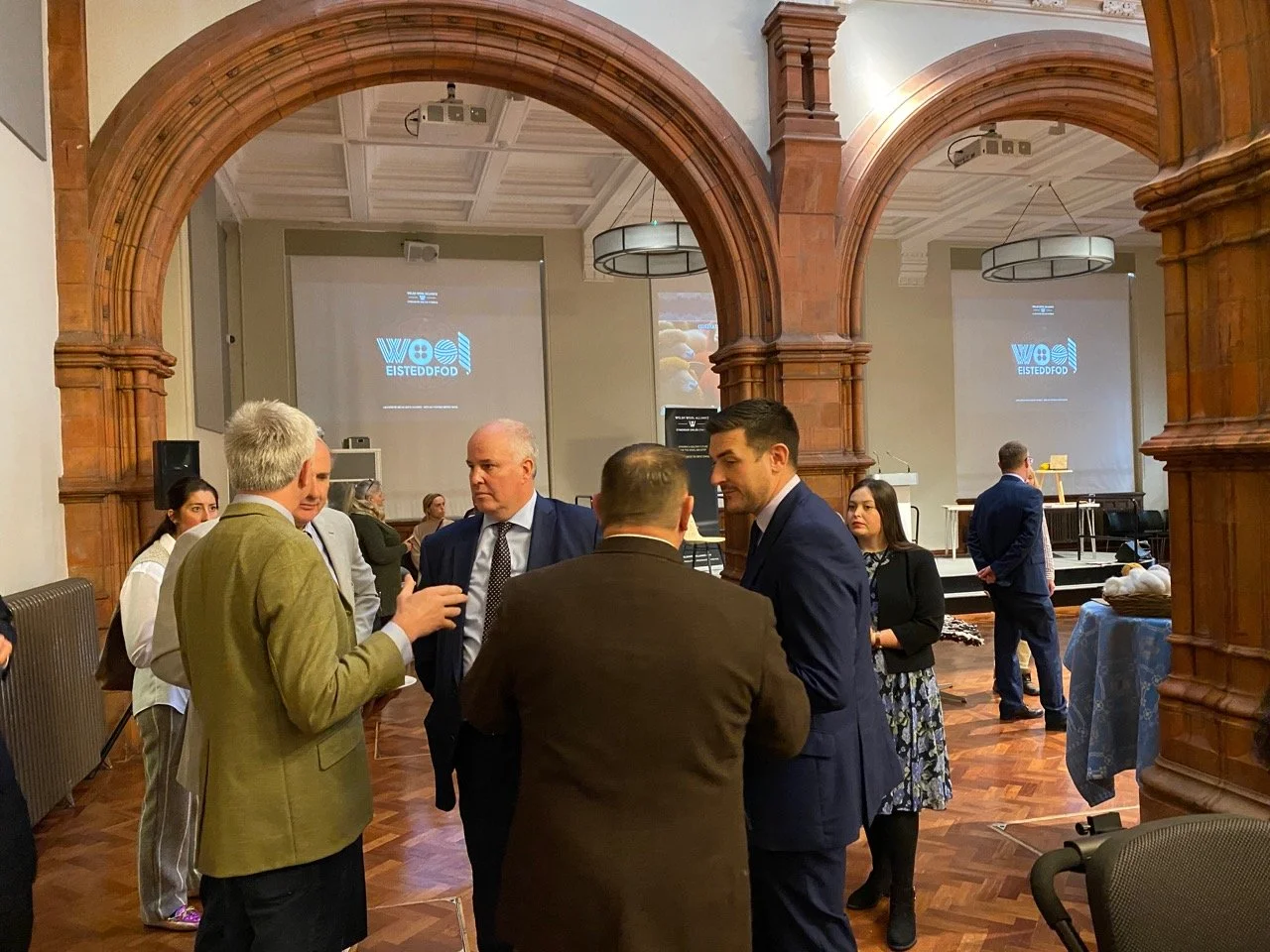 A group of people in formal attire engaging in conversation inside a grand, wood-paneled conference room with a high arched ceiling and projection screens displaying a logo and text.