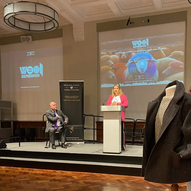 A woman in a pink blazer standing at a podium giving a presentation at an event for Eisteddfod, with a large projected image of sheep in the background. A man in a suit sits to her right on the stage, and a coat is hanging in the foreground.
