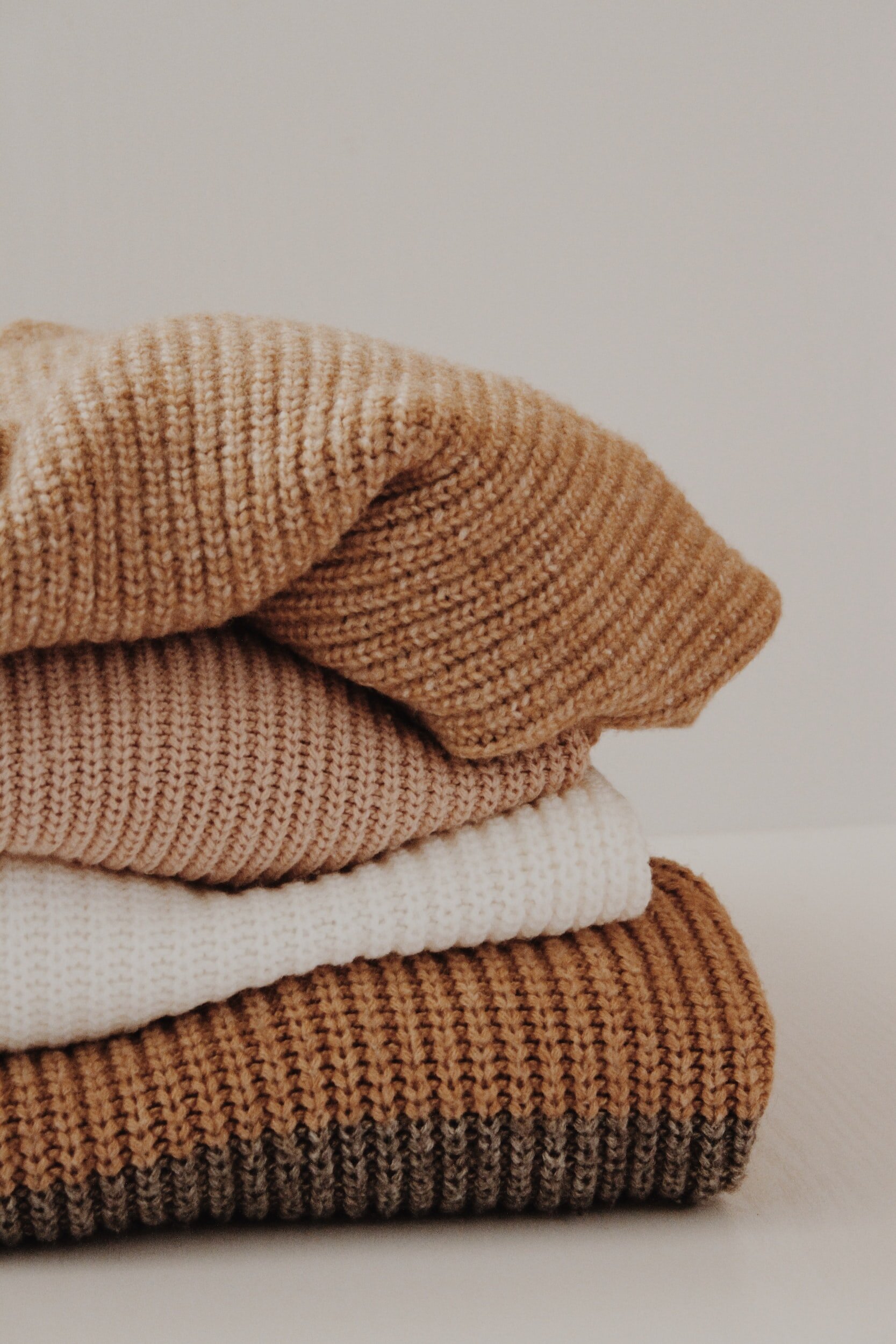 Stacked folded sweaters in neutral tones, including beige and white, on a plain background.