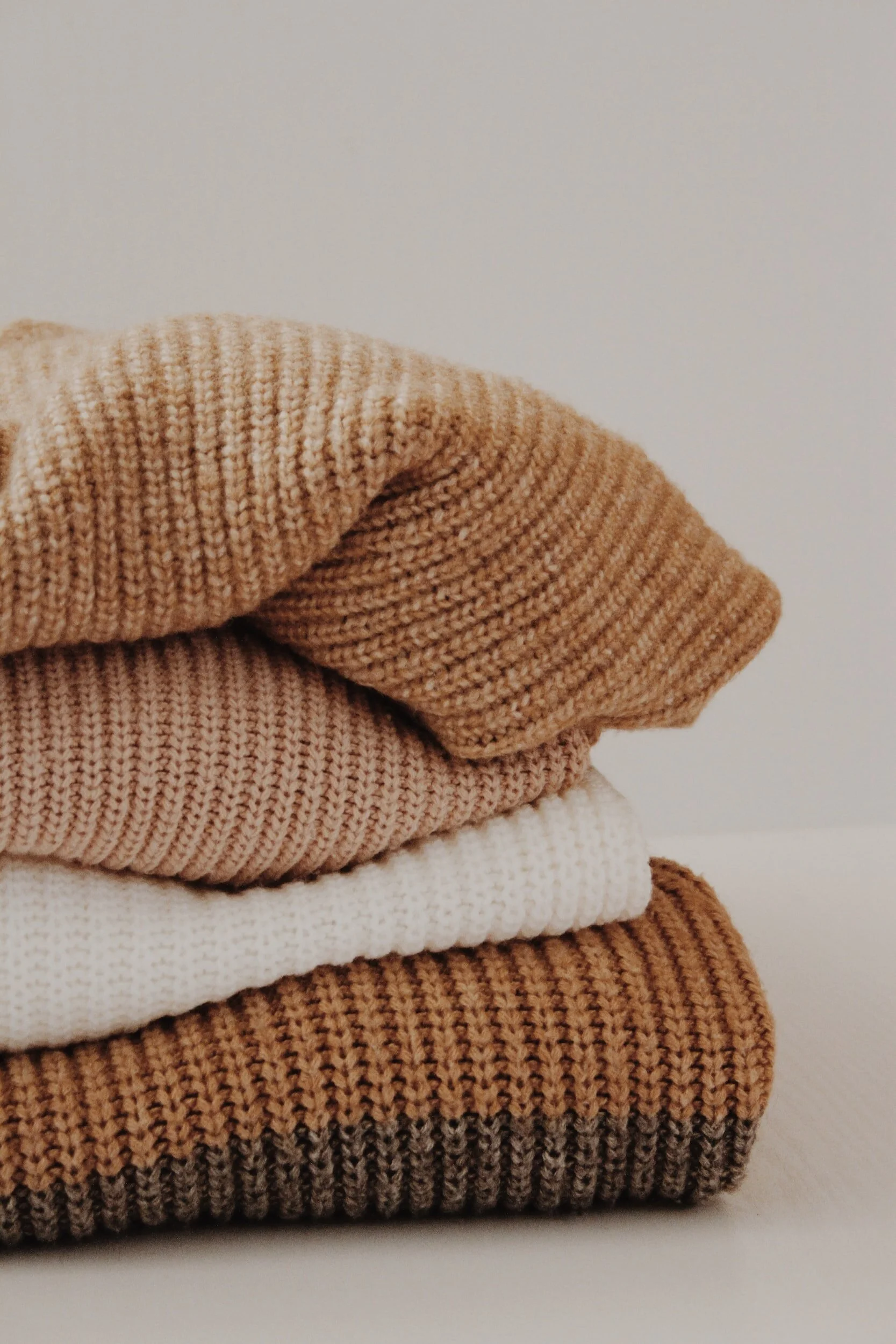 Stacked folded sweaters in neutral tones on a white surface against a plain background.