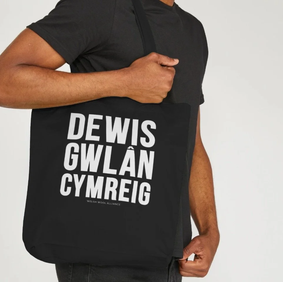 A person holding a black tote bag with the words 'DEWI SGWLÂN CYMREIG' printed in large white letters, and smaller text 'Welsh Wool Alliance' at the bottom.