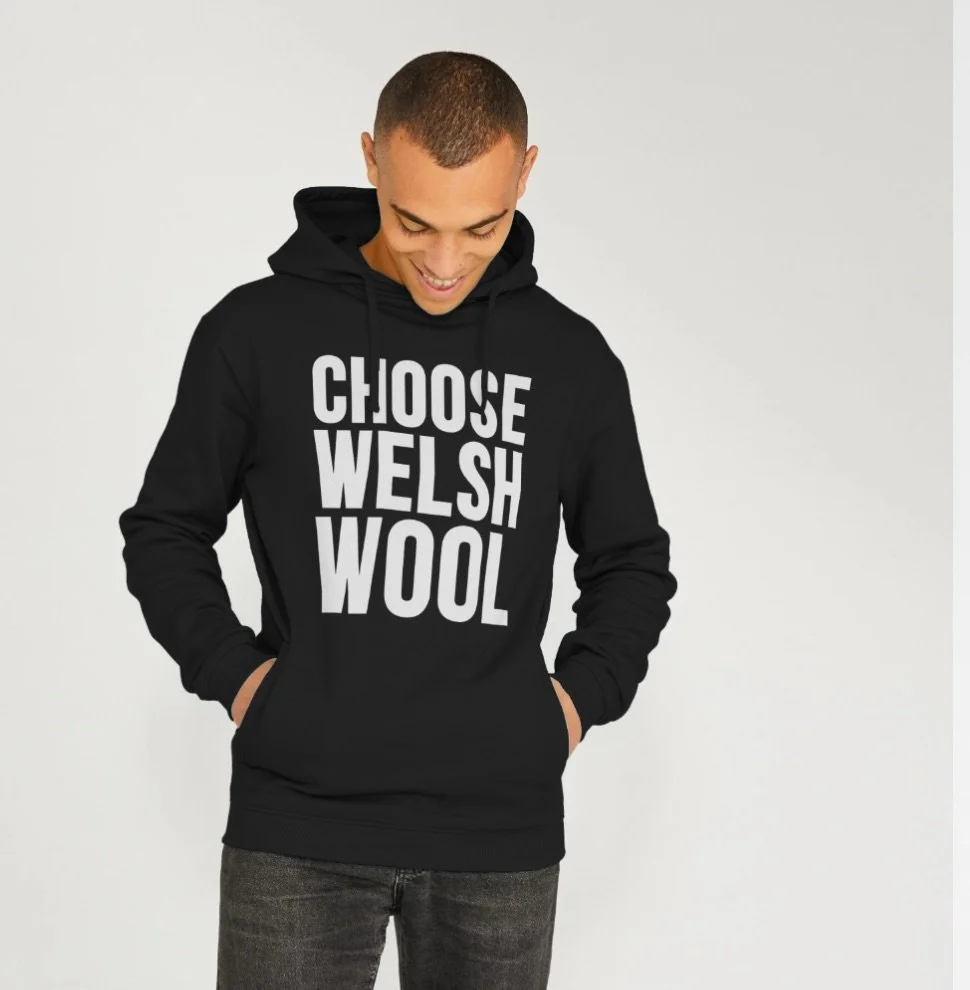 Young man wearing a black hoodie with bold white text that reads "CHOOSE WELSH WOOL". He is smiling and looking down, with hands in the hoodie pocket, standing against a plain white background.