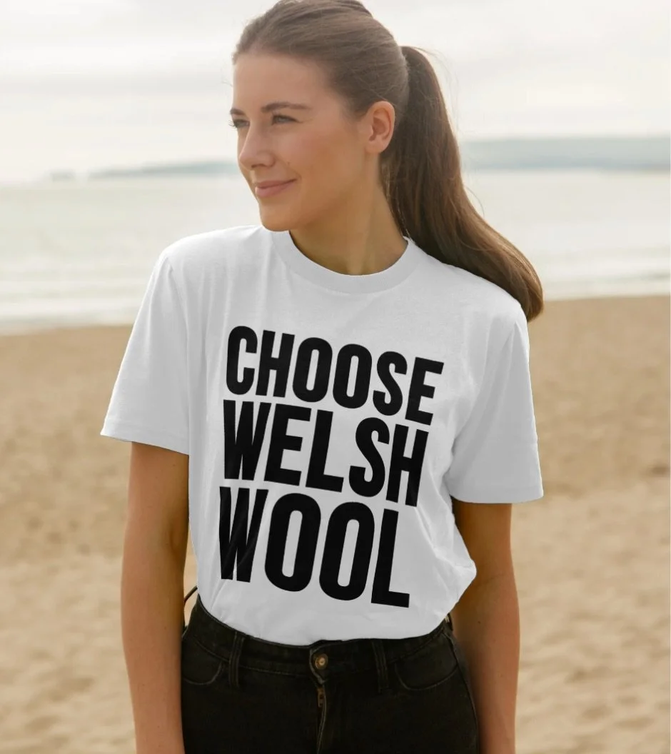 A woman standing on a beach, wearing a white T-shirt with bold black text that reads 'CHOOSE WELSH WOOL,' and black pants.