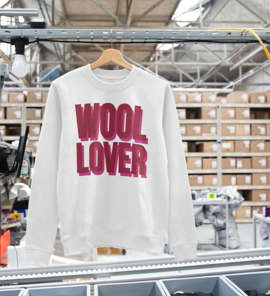 A white sweatshirt hanging on a wooden hanger on a metal rail in a warehouse. The sweatshirt has the phrase 'WOOL LOVER' printed in large, bold, pink letters on the front. Background contains shelves filled with cardboard boxes and warehouse items.
