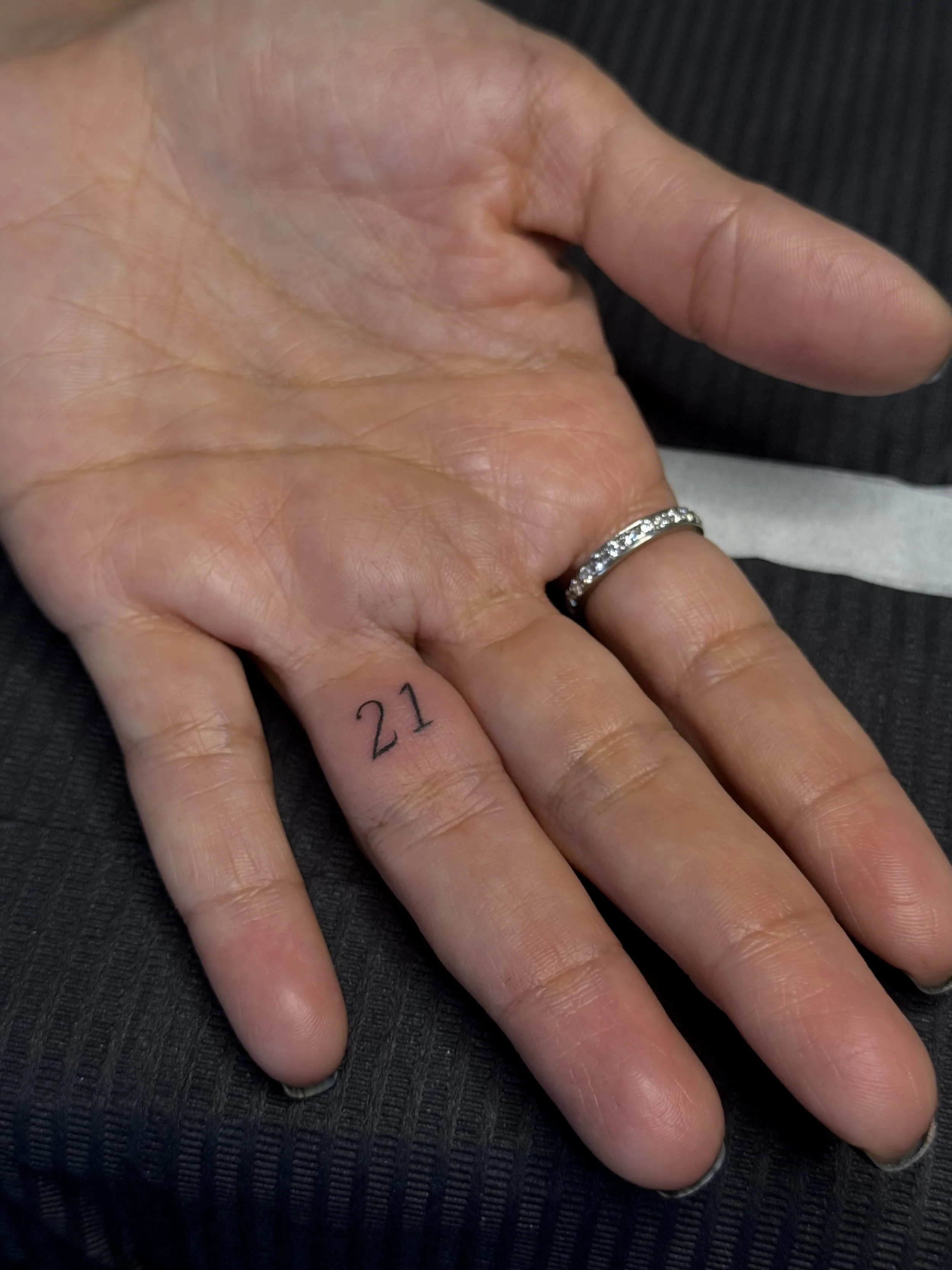 A person's hand with a silver ring that has embedded gemstones, resting on a dark textured surface. The person's index finger has the number '21' tattooed on it.