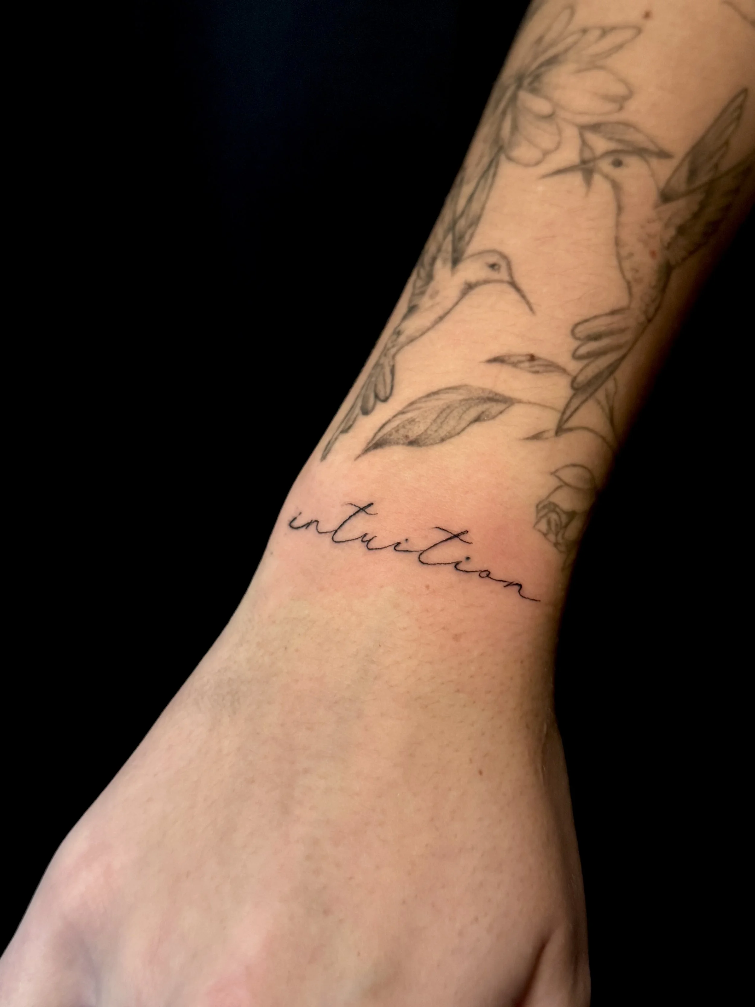 Close-up of a person's arm with a tattoo of a hummingbird and the word 'intuition' written in cursive.