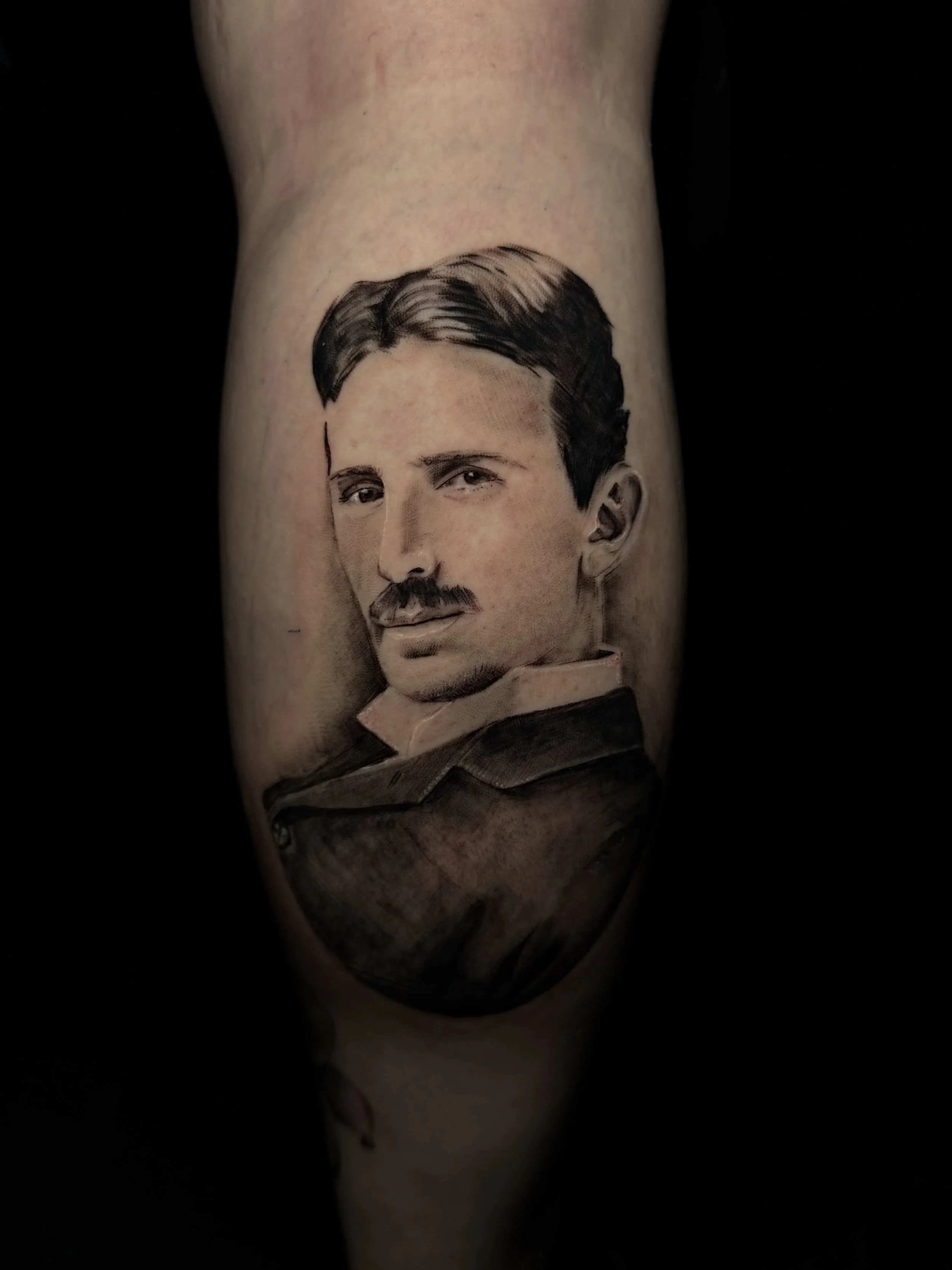 Tattoo of a man's face with dark hair and mustache on a person's arm, against a black background.