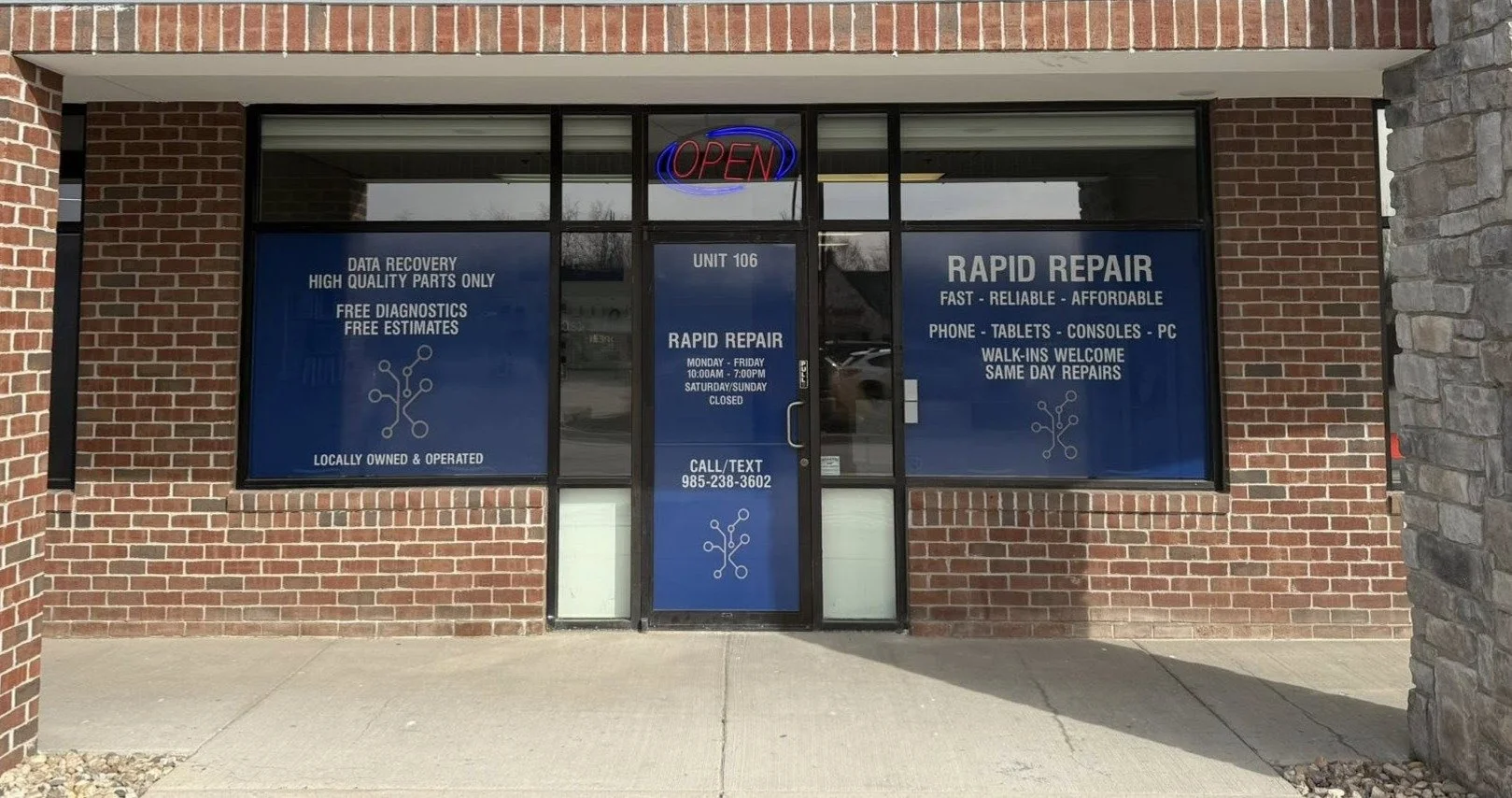 Storefront with blue windows advertising rapid repair services for electronics, open sign, brick facade, and concrete sidewalk.