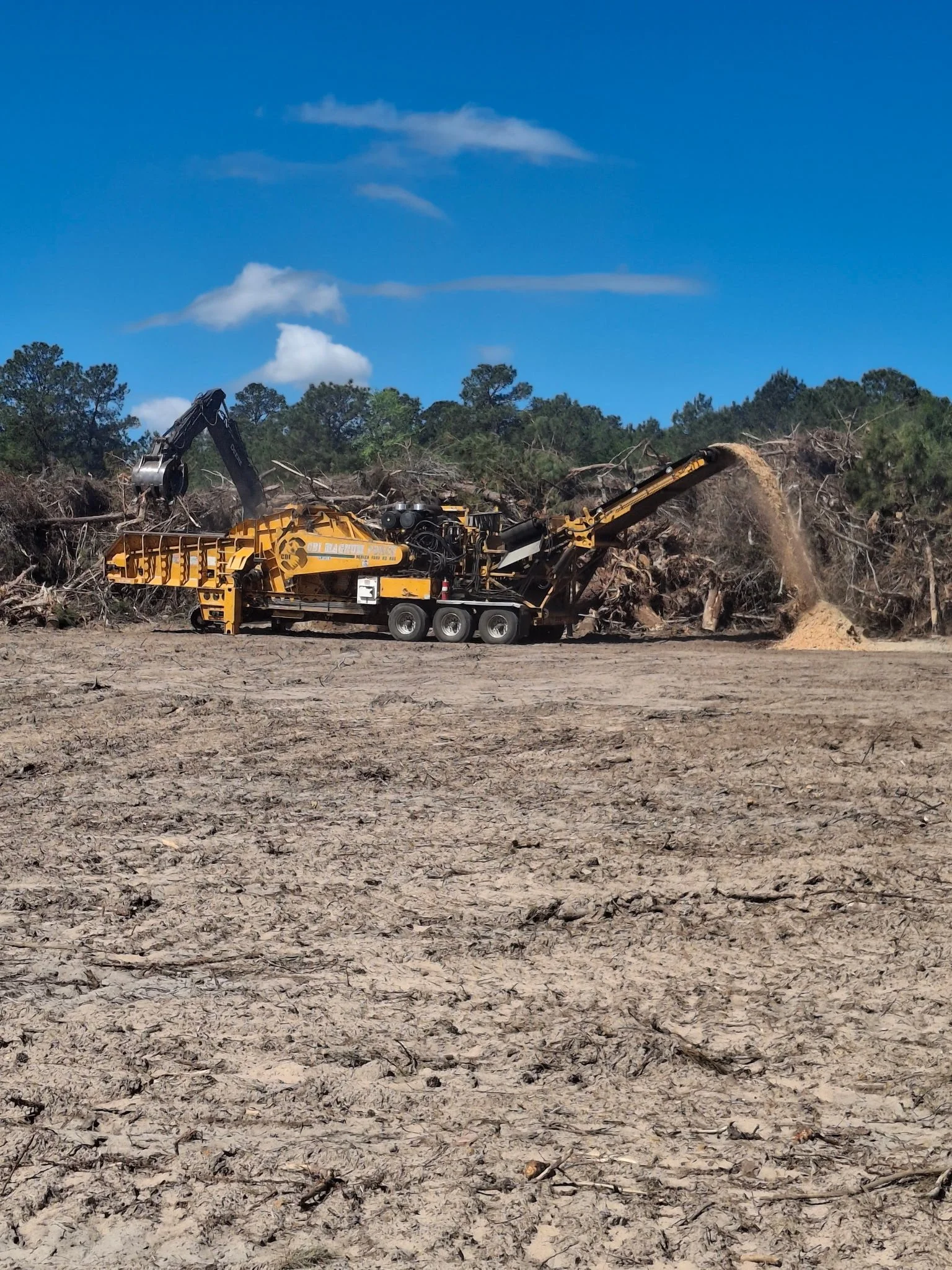 A large yellow grinding machine on wheels operates on a barren, cracked dirt field, processing a pile of branches and wood debris. In the background, there are trees and a partly cloudy blue sky.