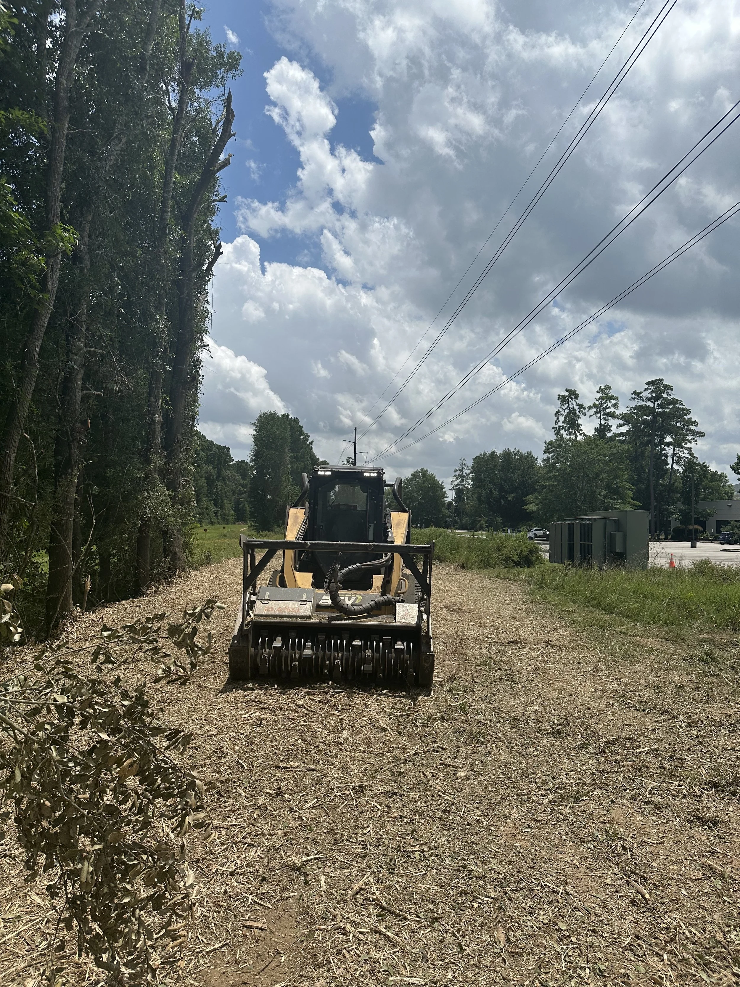 A construction or landscaping vehicle on a dirt path next to a tree-lined area under a partly cloudy sky.