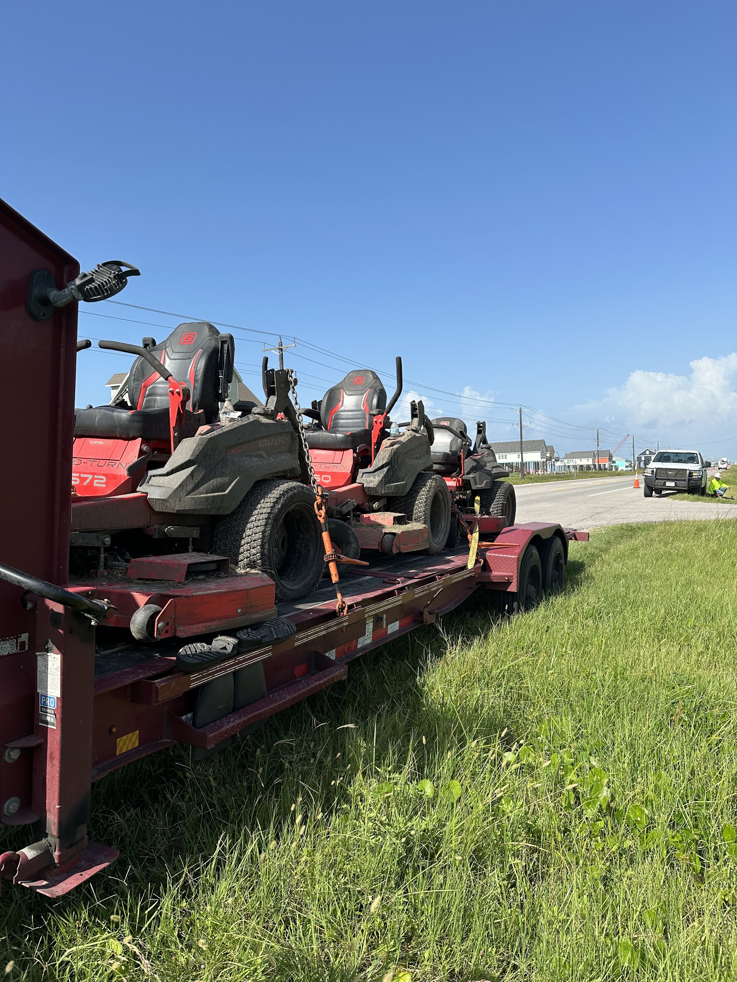 Red flatbed trailer carrying three riding lawn mowers parked on grass beside a road, with traffic cones and vehicles in the background under a bright blue sky.