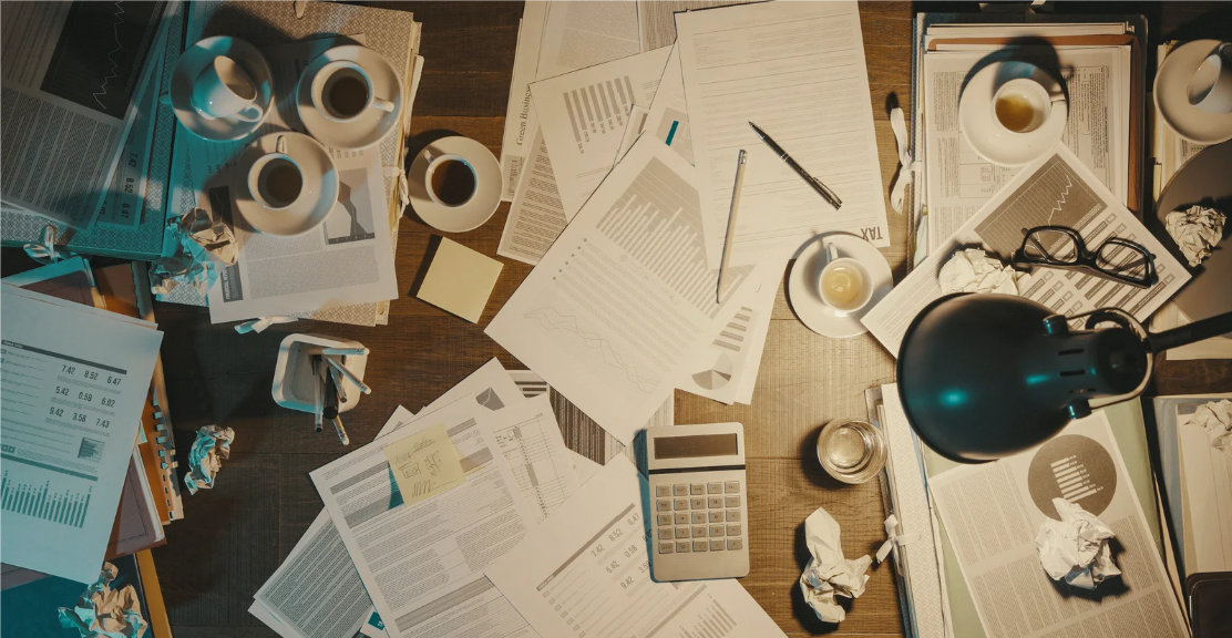 Cluttered desk with papers, charts, a calculator, glasses, coffee cups, a lamp, and crumpled papers.
