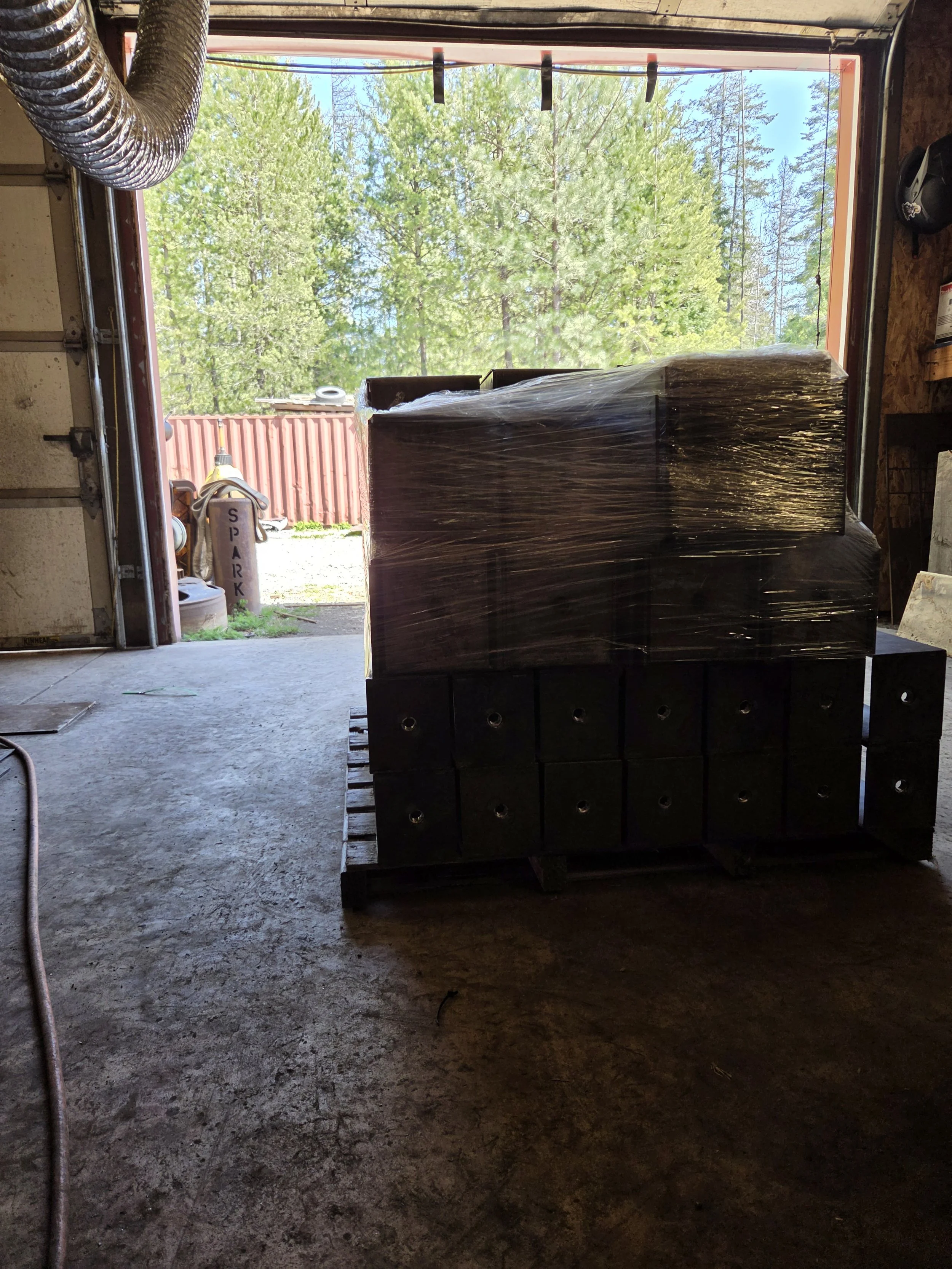 View from inside a garage showing a pallet stacked with wrapped items outside, with trees and blue sky in the background.