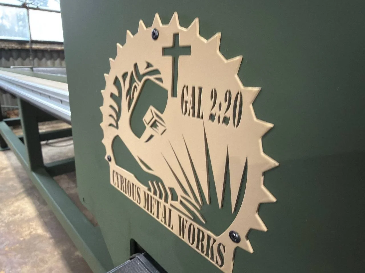Metal sign with a circular gear design, a cross, a sunflower, and text reading "GAL 2:20" and "CURIOS METAL WORKS".