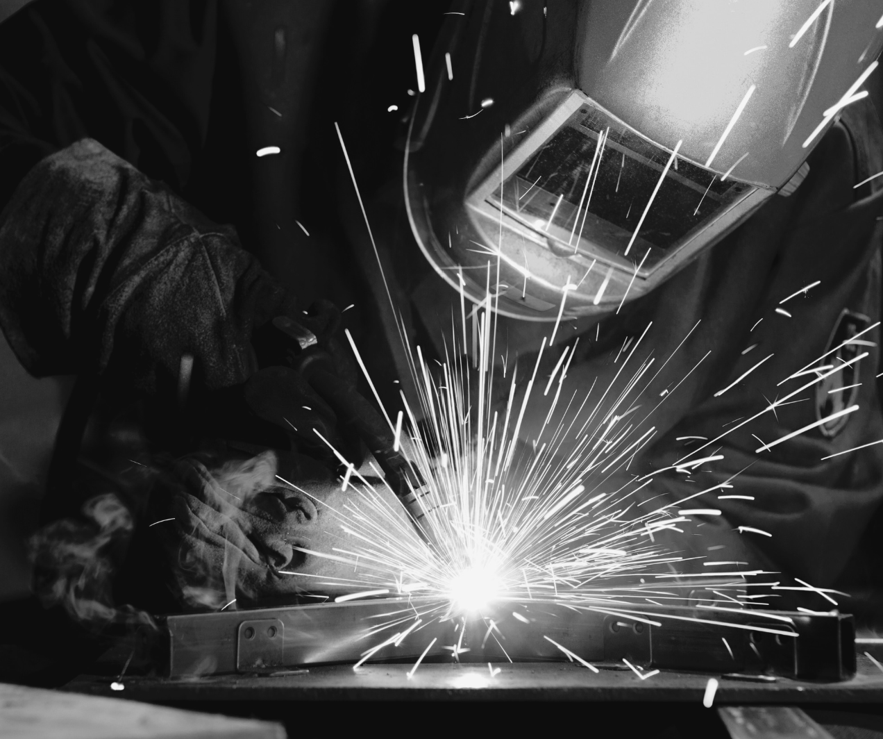 A person welding metal, sparks flying, wearing protective gear including a welding helmet and gloves.