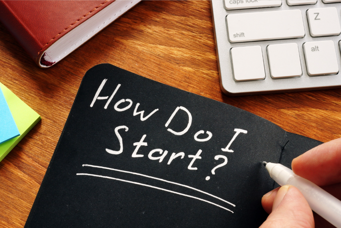 A person writing 'How Do I Start?' on a black chalkboard with a white marker, surrounded by a white keyboard, a red notebook, and colorful sticky notes on a wooden desk.