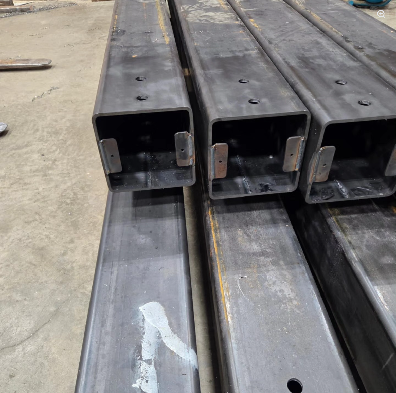 Stacked hollow steel rectangular tubes with welded brackets, placed on a concrete floor in an industrial setting.