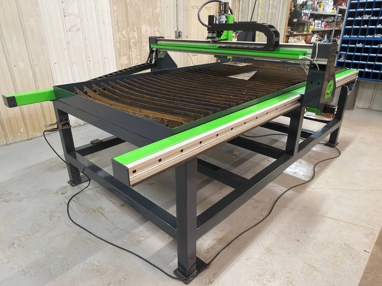 A large green and black CNC router machine in a workshop with tools and storage shelves in the background.