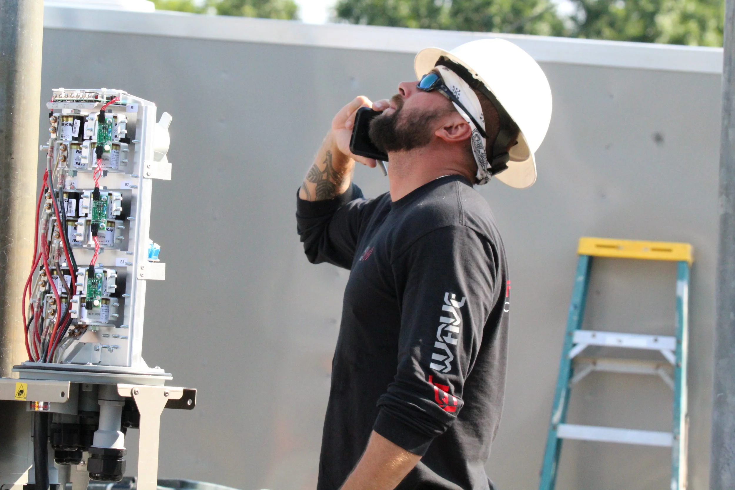 A man wearing a construction helmet and sunglasses talking on a cellphone while working on an electrical panel outdoors, with a ladder in the background.