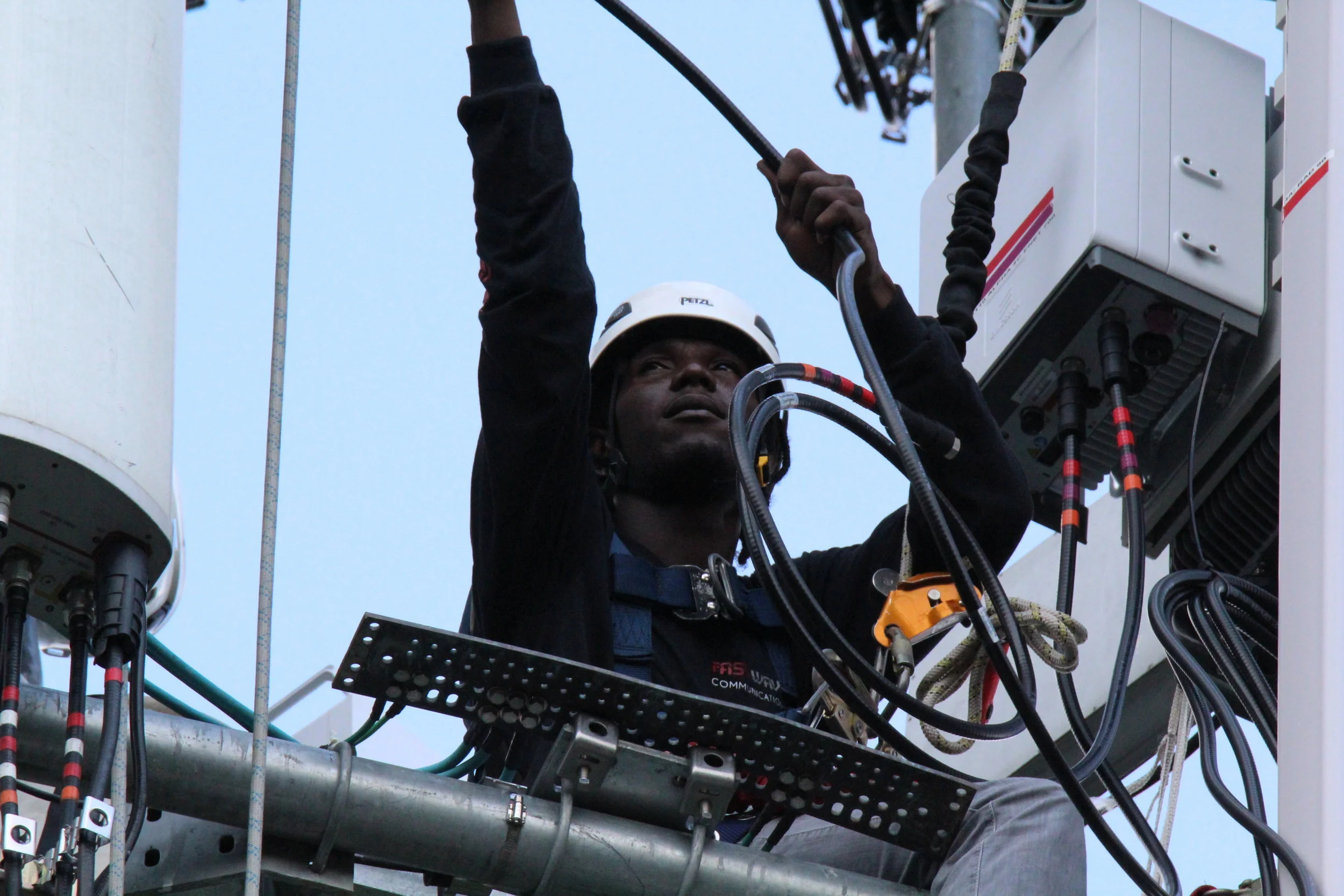 A technician wearing a helmet working on a communication tower, surrounded by wires and equipment.