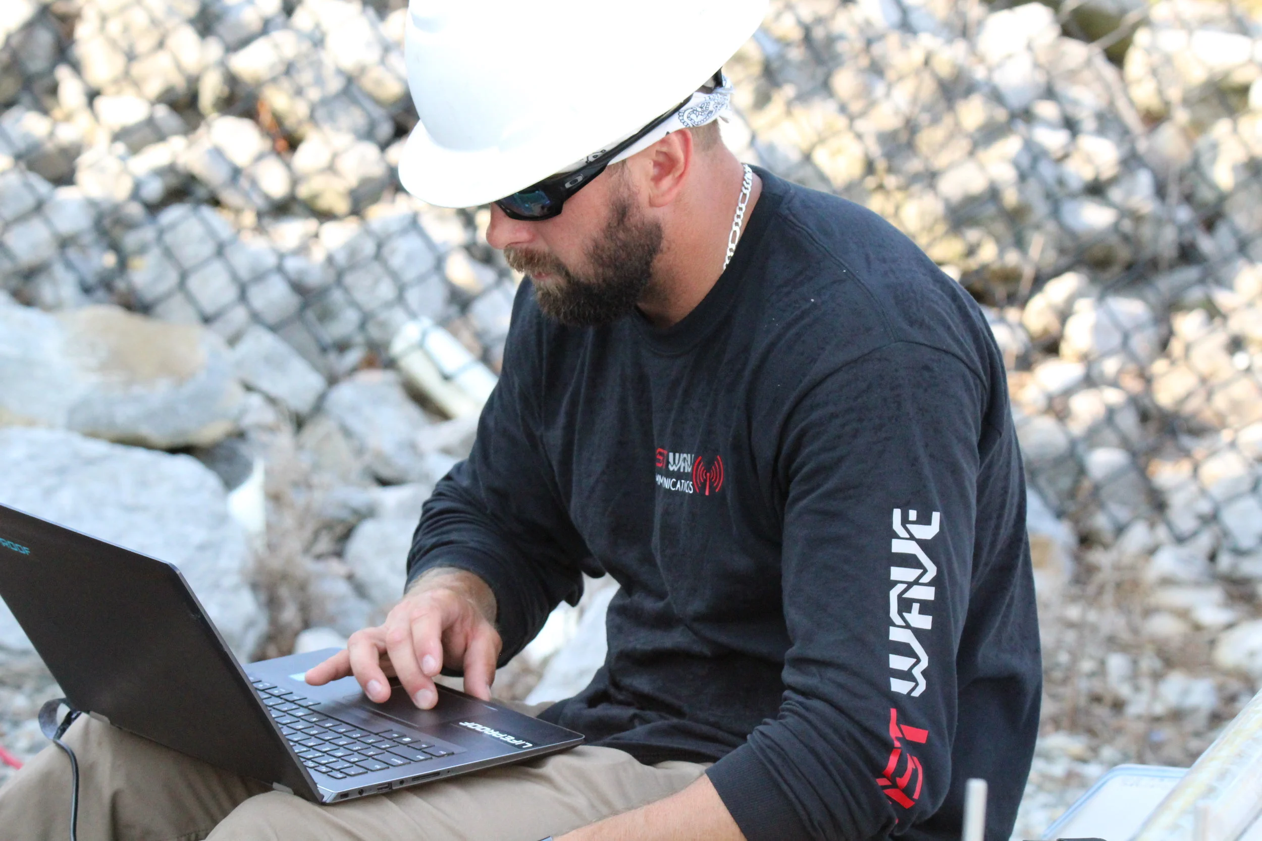 Man wearing a white hard hat, sunglasses, and a black long-sleeve shirt working on a laptop outdoors with a rocky background.