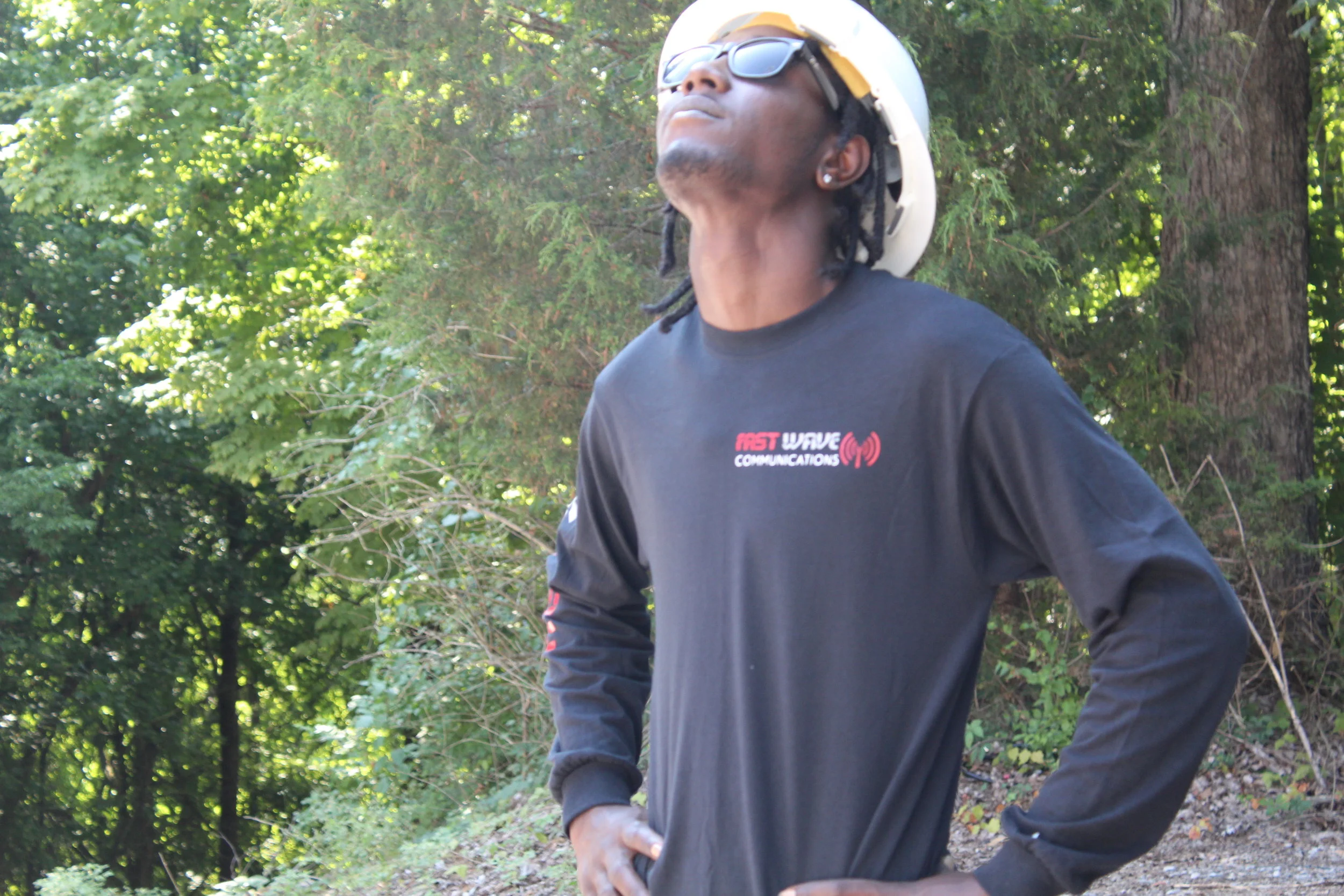 A young man standing outdoors with his head tilted back, wearing sunglasses, a black long-sleeve shirt, and a white helmet, surrounded by green trees.