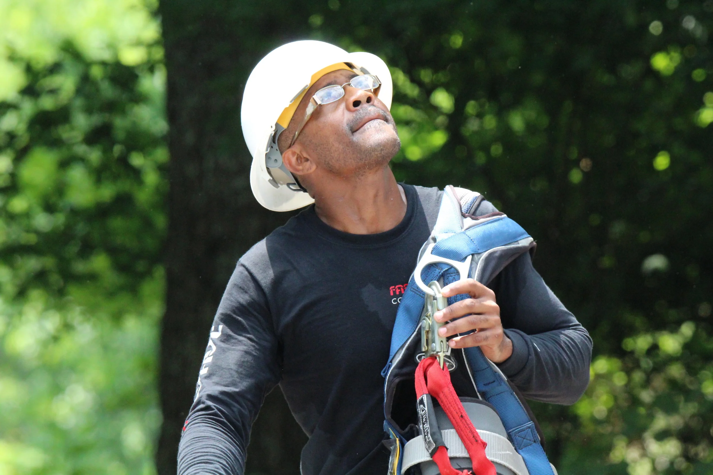 A man wearing a white safety helmet, glasses, and a black shirt is holding a climbing harness while standing outdoors in a green, forested area.