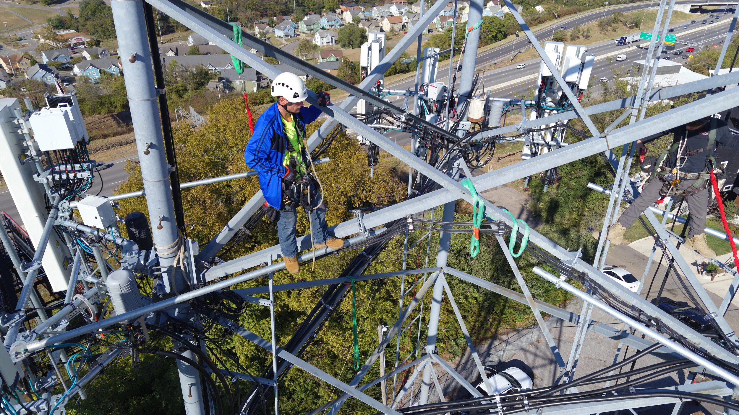 Two technicians working on top of a tall communication tower with various cables and equipment, overlooking a suburban area and highways.