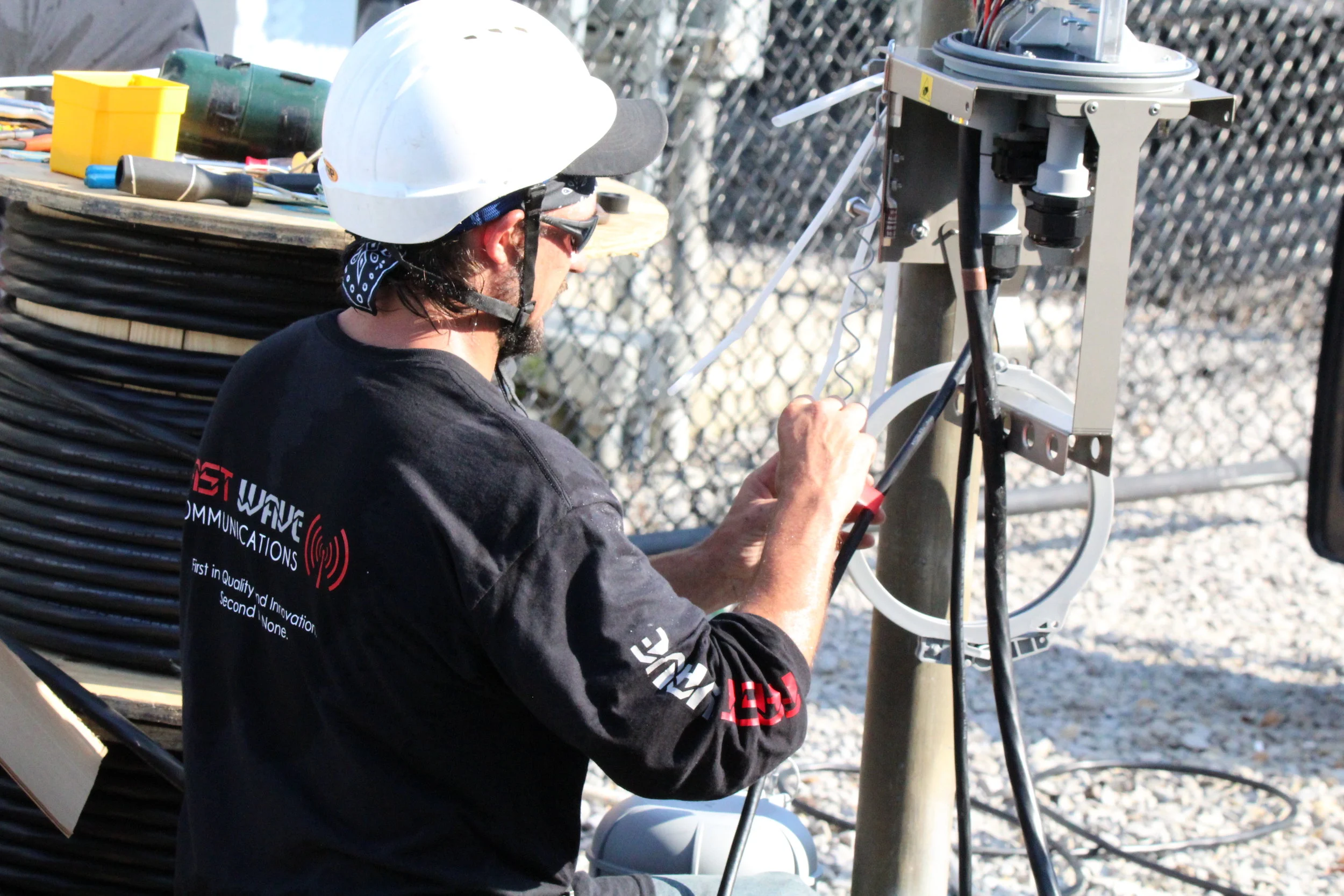 A technician working outdoors on a communication or electrical device mounted on a pole, wearing a white hard hat, sunglasses, and a black shirt with red and white text.