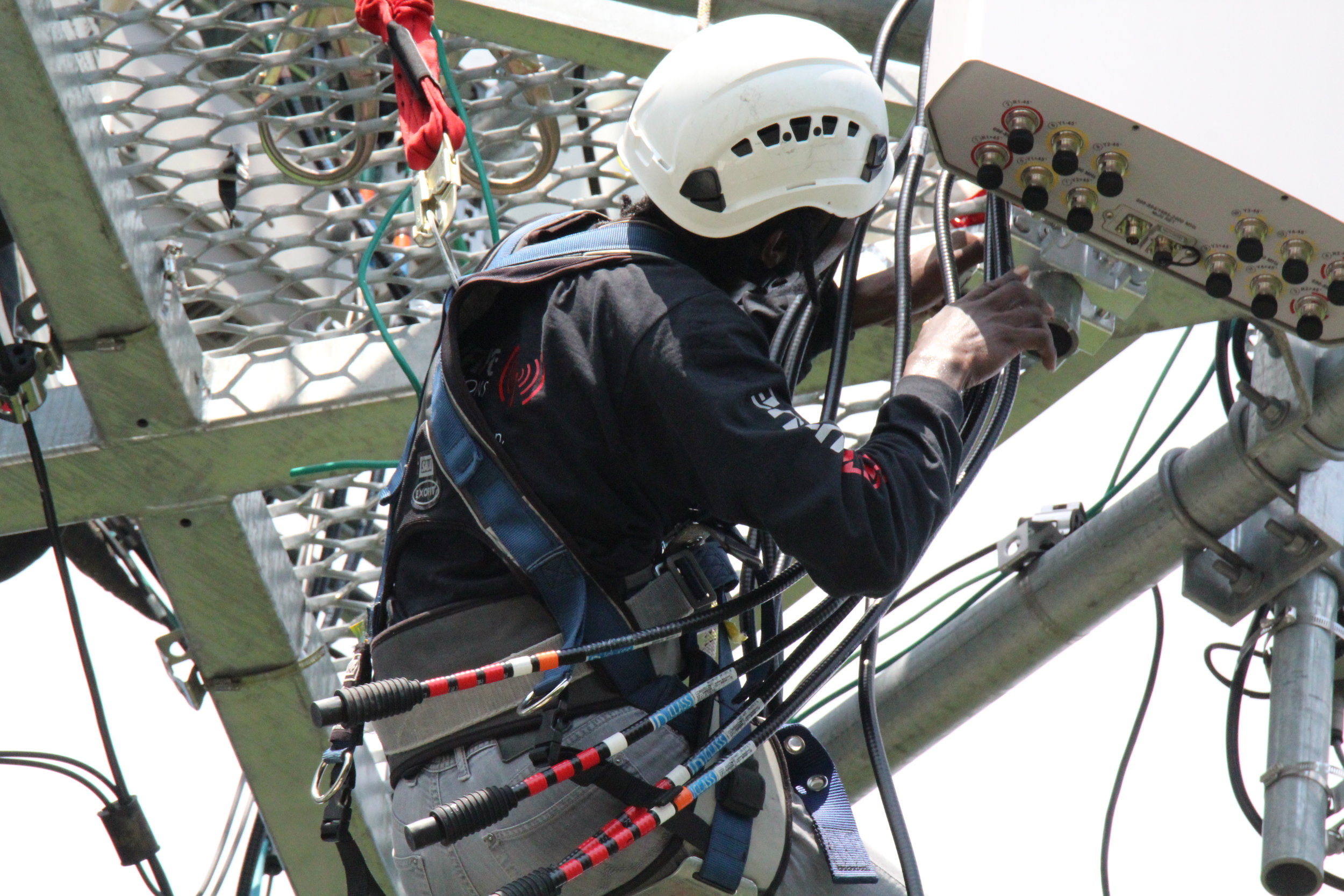 A technician wearing a safety helmet and harness working on electrical equipment on a metal structure.