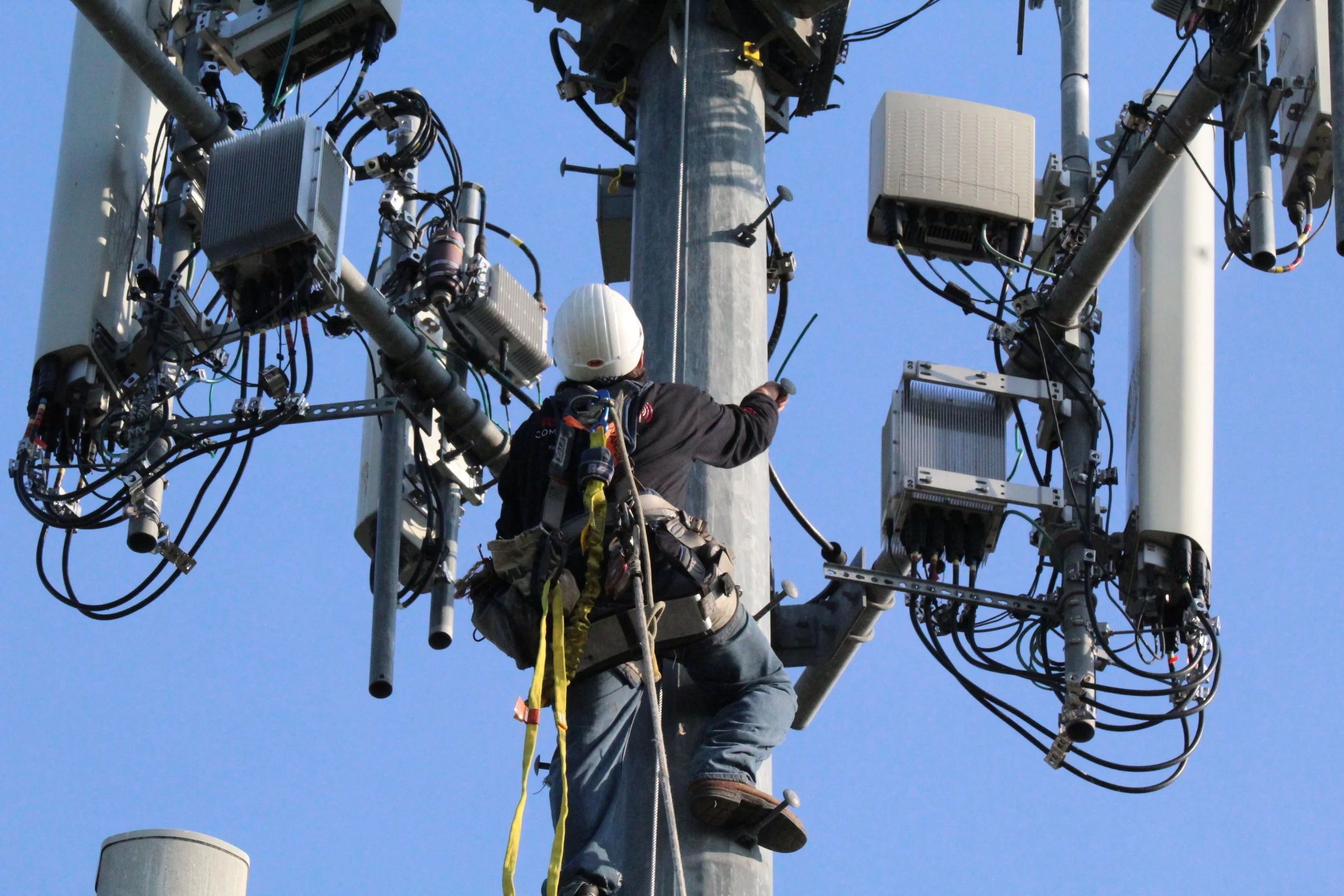 A technician is working on a utility pole fitted with various electrical and communication equipment against a clear blue sky.