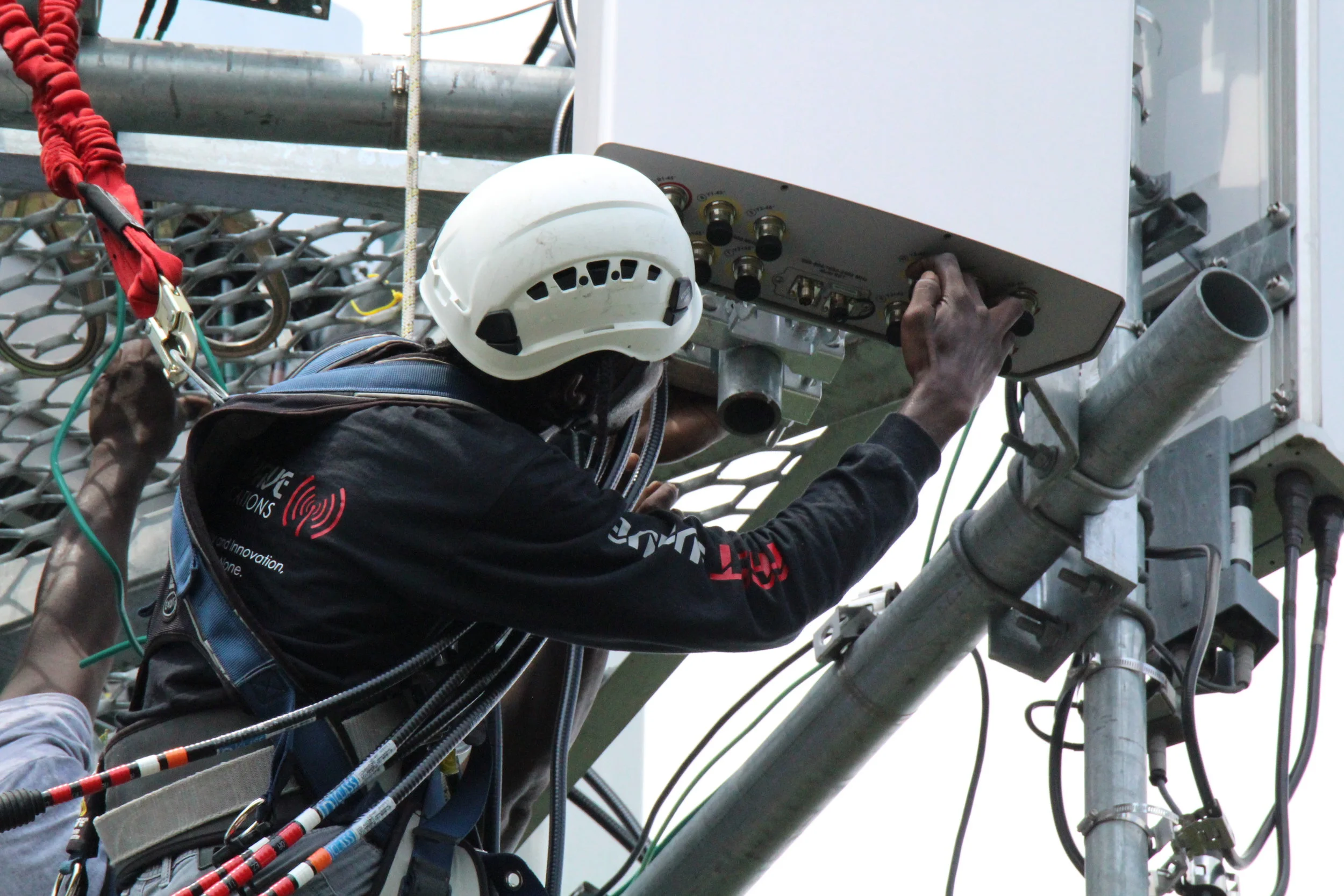A worker wearing a white safety helmet and black long sleeve shirt with red and white lettering is adjusting a control panel on electrical equipment mounted on a metal pole, with tangled wires and pipes around.