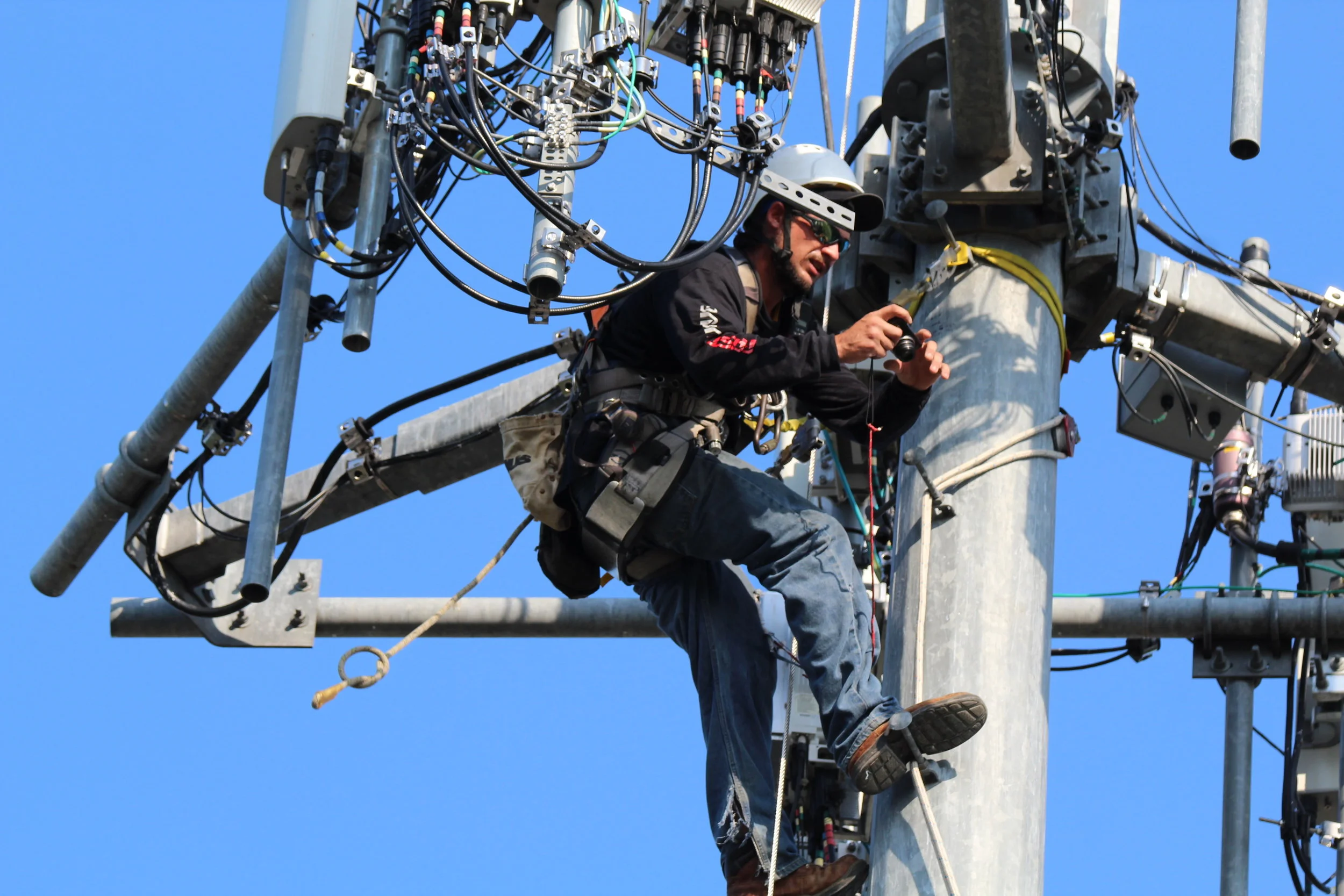 A technician wearing a safety helmet and harness is working on an electrical power pole, inspecting or repairing equipment, with a clear blue sky in the background.