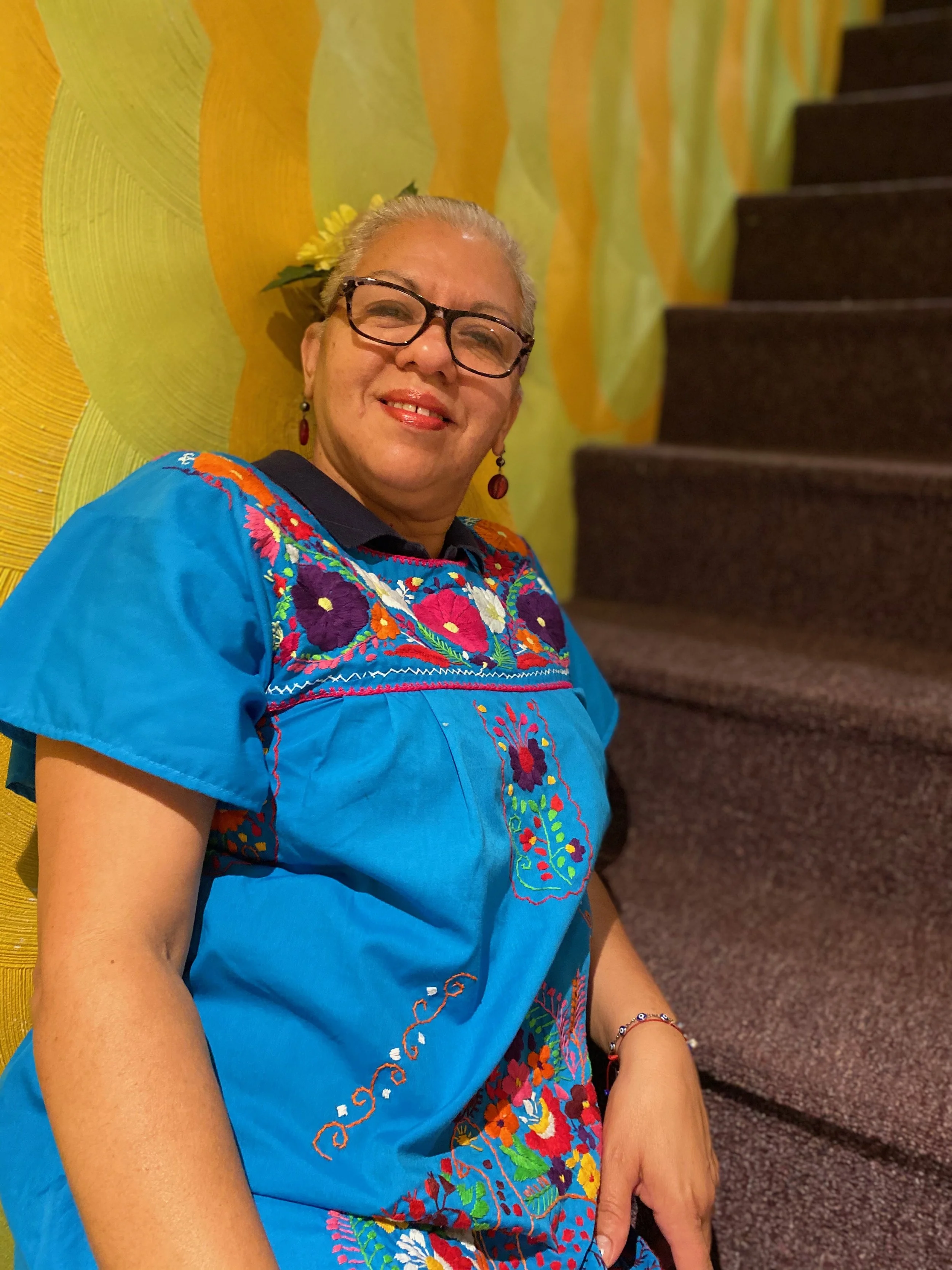 Mujer mayor con gafas, vestido azul con bordados coloridos, pendientes rojos y pulsera, sonriendo en un fondo decorado con paredes pintadas en colores amarillo y verde, y escaleras de color marrón.