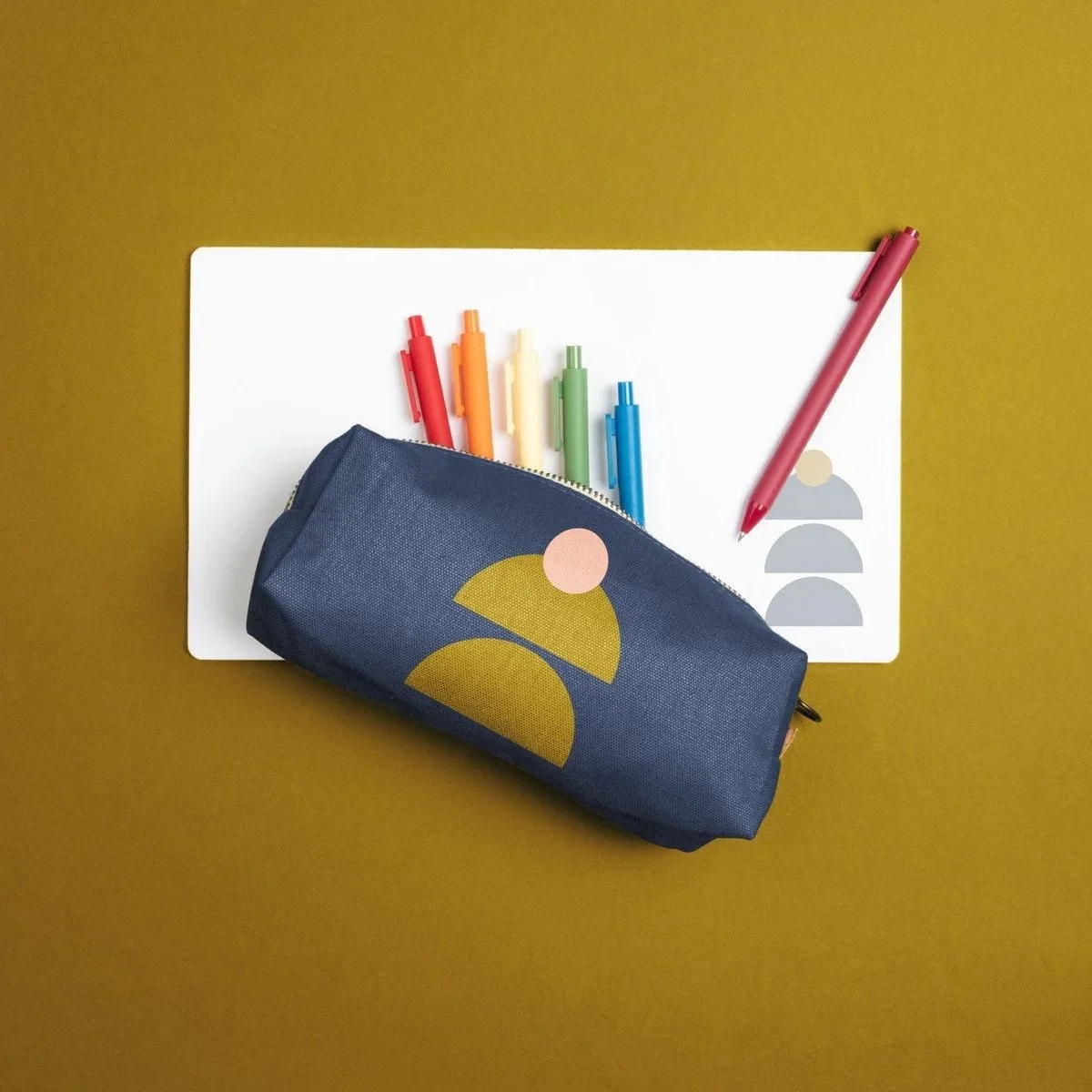 A flat lay of colorful pens, a navy pencil case with geometric design, a white sheet of paper, and a red pen on a yellowish background.